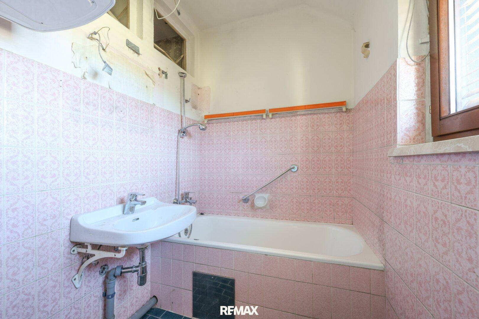 Badezimmer mit rosa Fliesen, Badewanne, Waschbecken und Haltegriffen, renovierungsbedürftig.