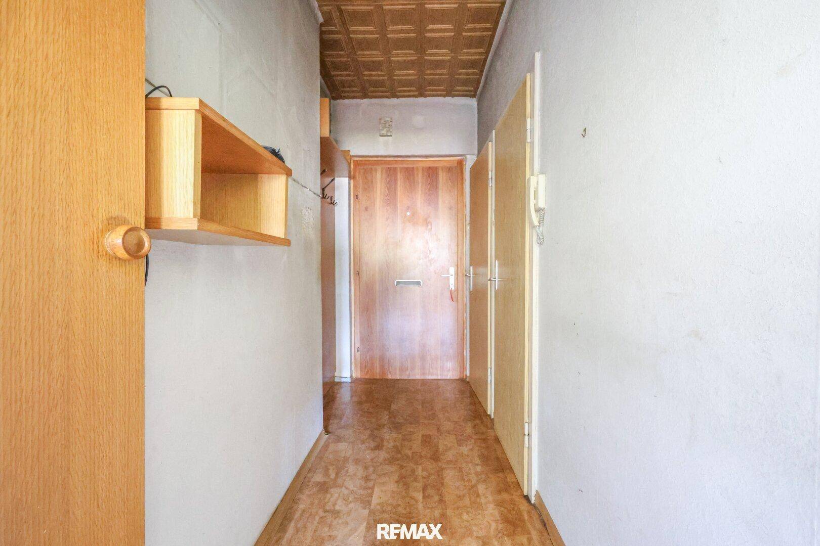 Langer Flur mit Holztür, Holzregal und gemusterter Deckenverkleidung in originalem Zustand.