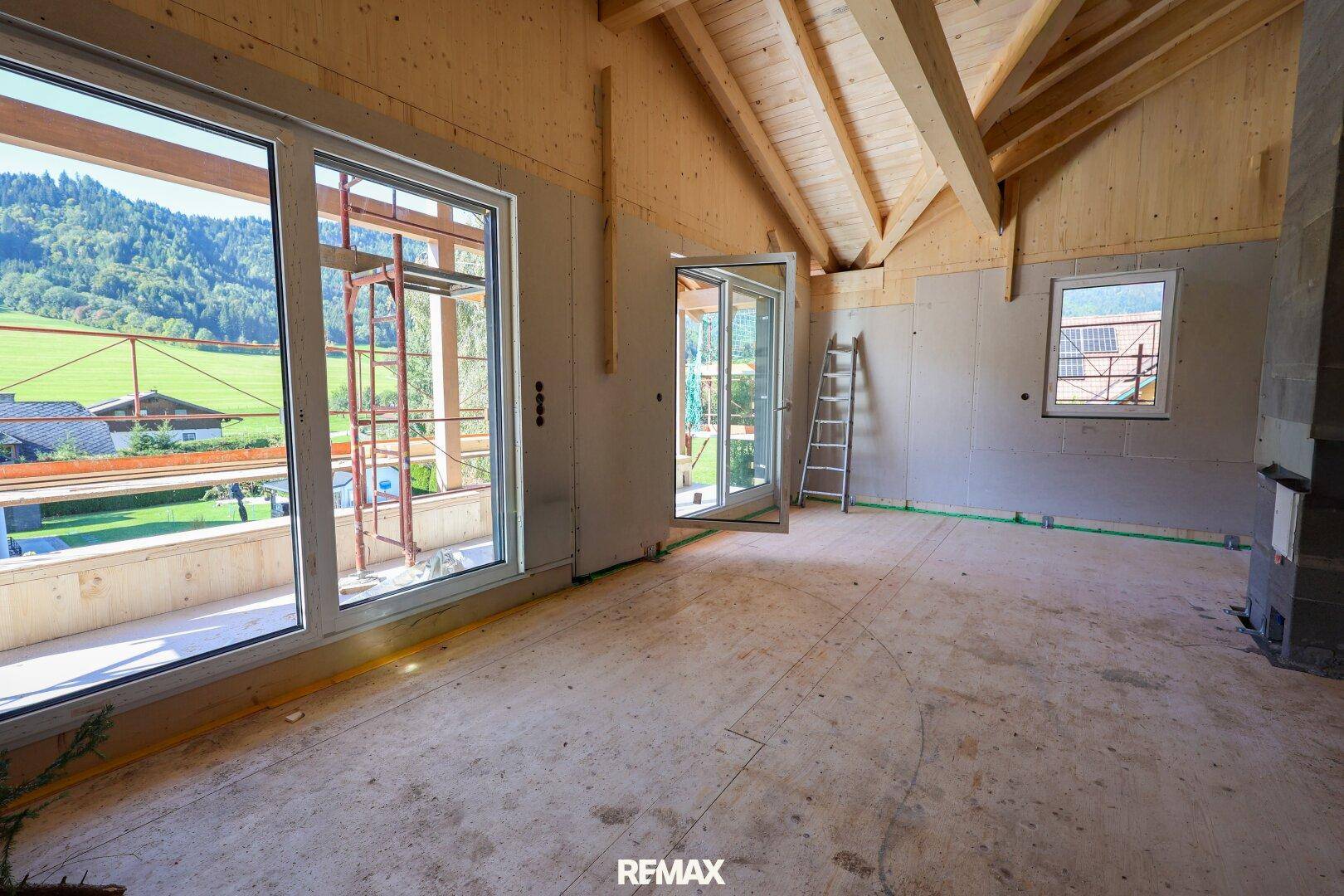 Heller Innenraum im Rohbau mit großen Fenstern, Holzbalken und Blick ins Grüne.