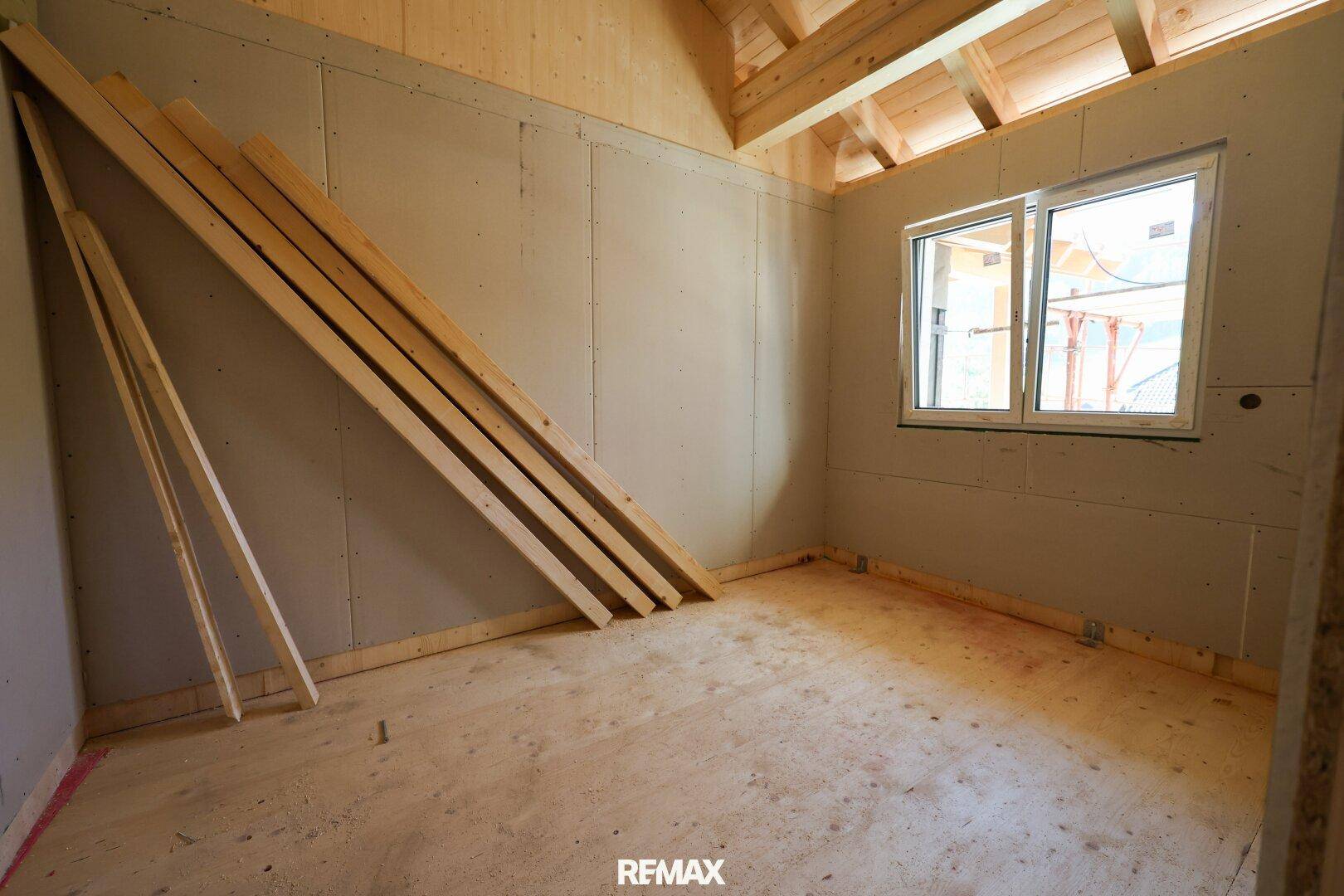 Unfertiger Raum mit Holzbalken, Gipskartonwänden und einem Fenster mit Blick nach draußen.