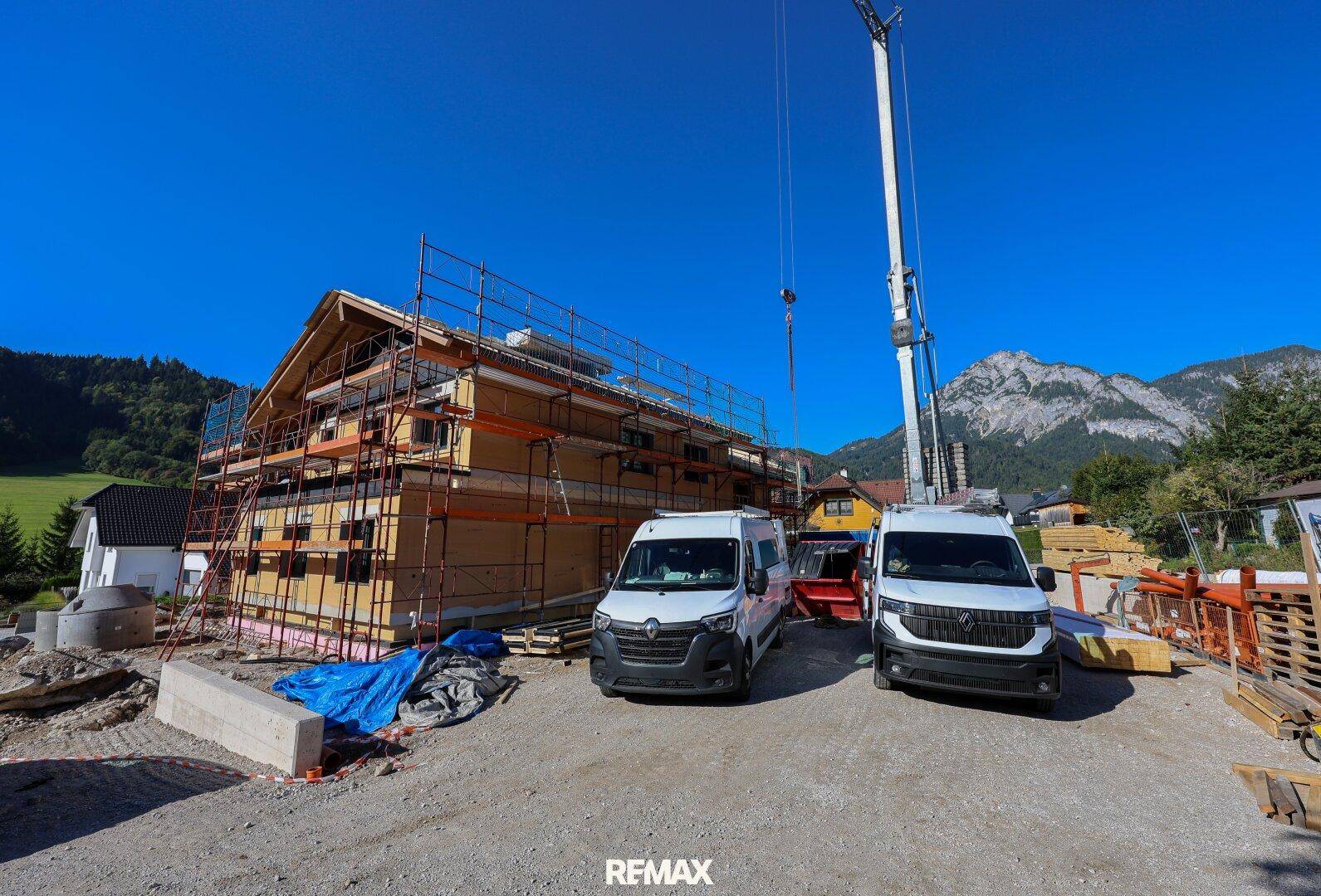 Baustelle eines Neubaus mit Gerüst, Kran und Lieferwagen, eingebettet in eine ländliche Umgebung.