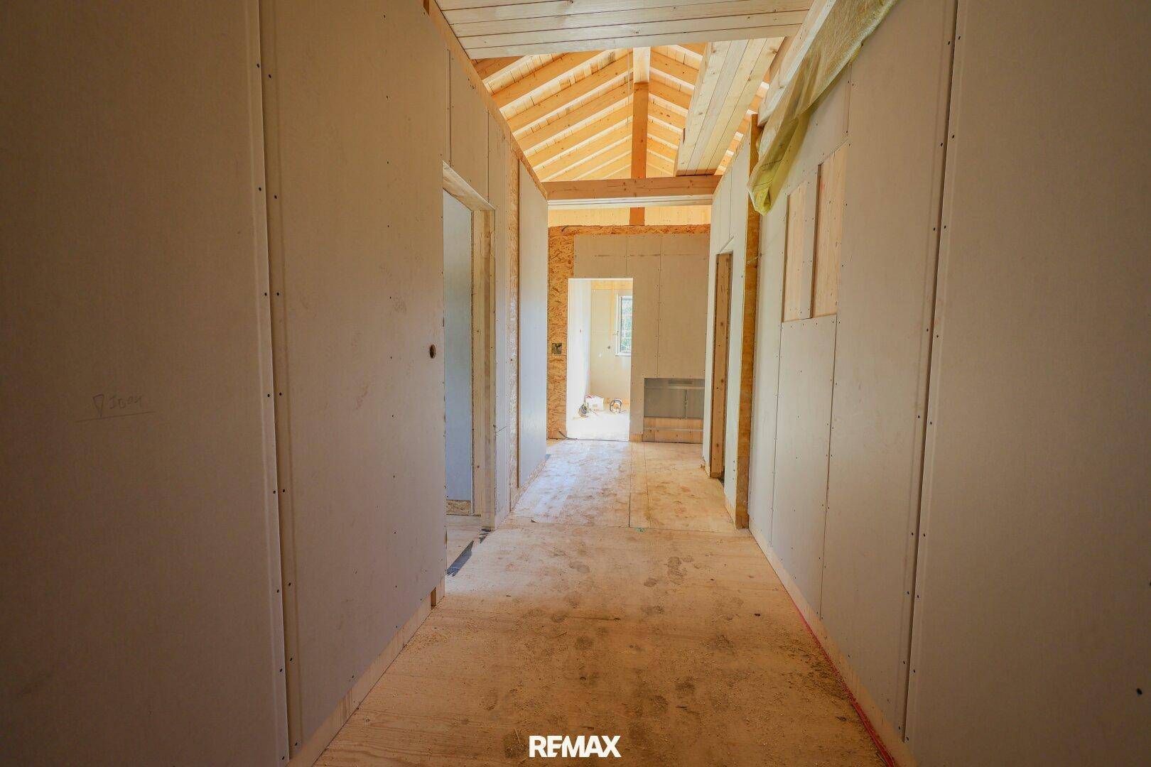 Flur im Rohbau mit sichtbarer Holzkonstruktion und unfertigen Wänden, der zu weiteren Räumen führt.
