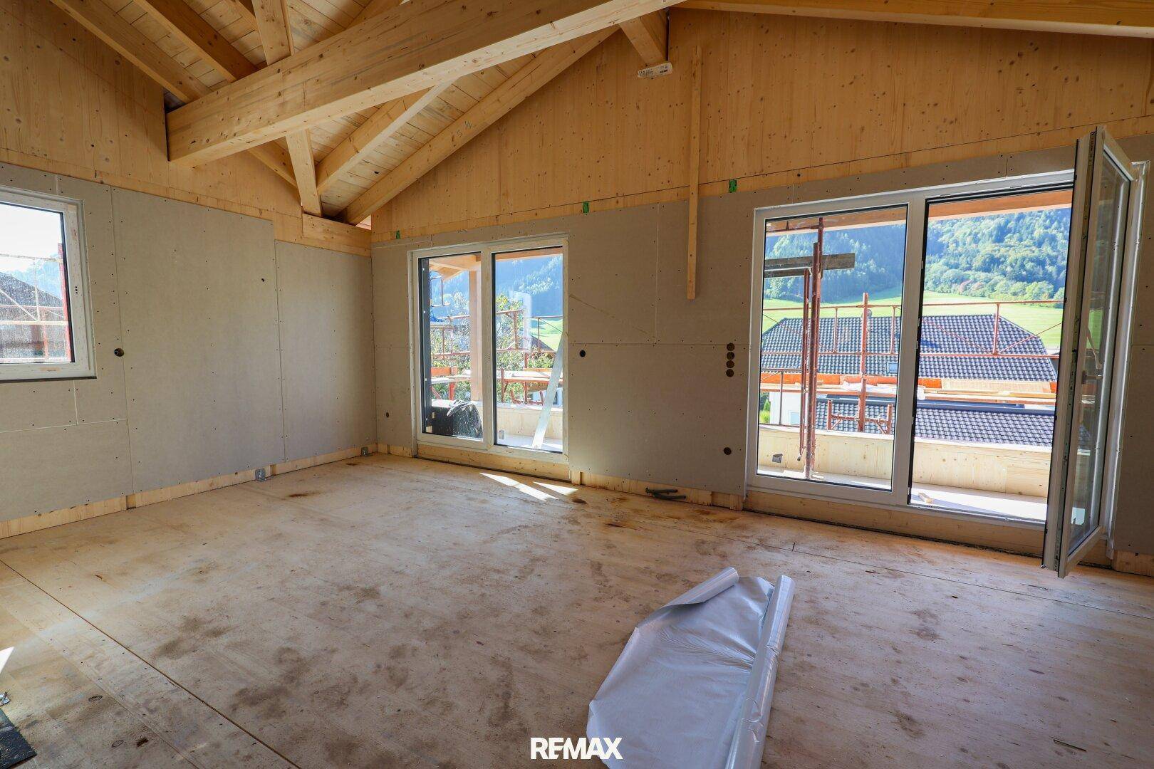 Geräumiger Wohnbereich im Rohbau mit sichtbarer Holzkonstruktion und großen Fenstern mit Bergblick.