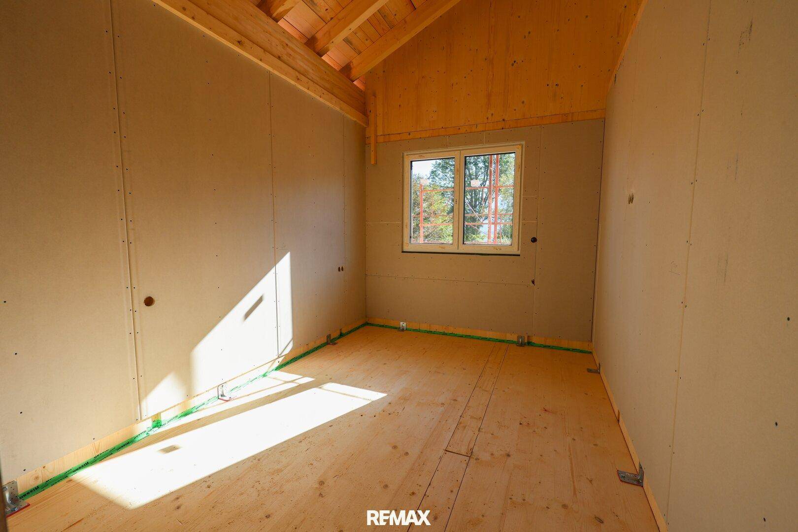 Schlafzimmer im Rohbau mit Holzboden und einem Fenster, das viel natürliches Licht hereinlässt.