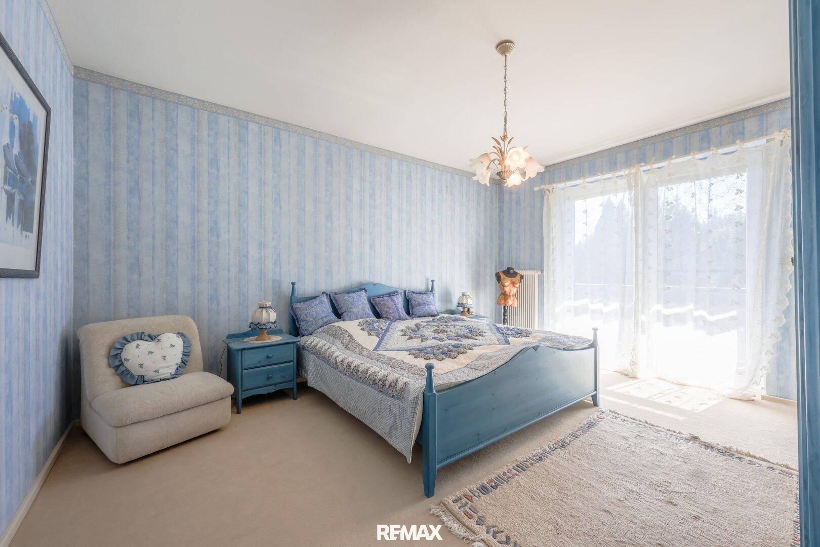 Schlafzimmer mit blau gestreifter Tapete, Doppelbett und Zugang zu einem Balkon.