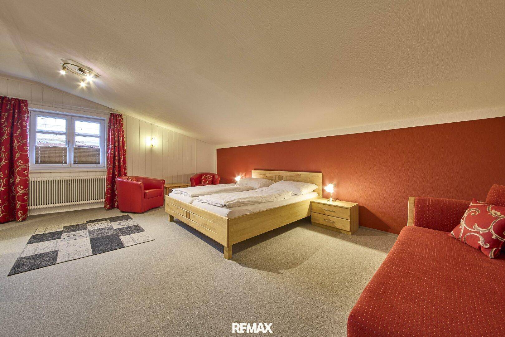 Geräumiges Schlafzimmer im Dachgeschoss mit Holzbett, roter Akzentwand und Teppichboden.