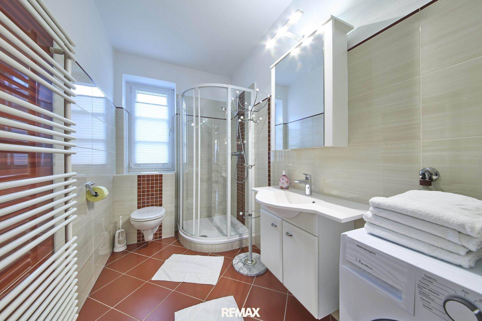 Modernes Badezimmer mit Dusche, Waschmaschine und beheiztem Handtuchhalter in warmen Farbtönen.
