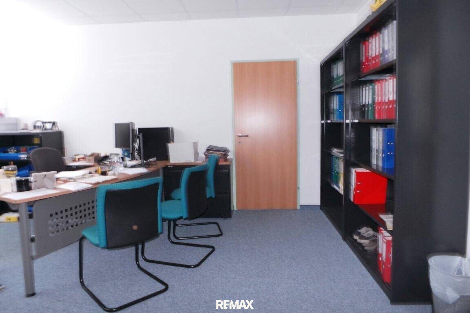 Ein funktionales Büro mit blauem Teppichboden, Schreibtischen und einem großen schwarzen Bücherregal.
