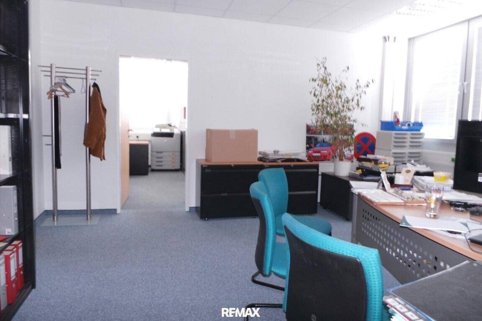 Heller Büroraum mit blauem Teppichboden, Schreibtischen und einem Durchgang zu einem weiteren Arbeitsbereich.