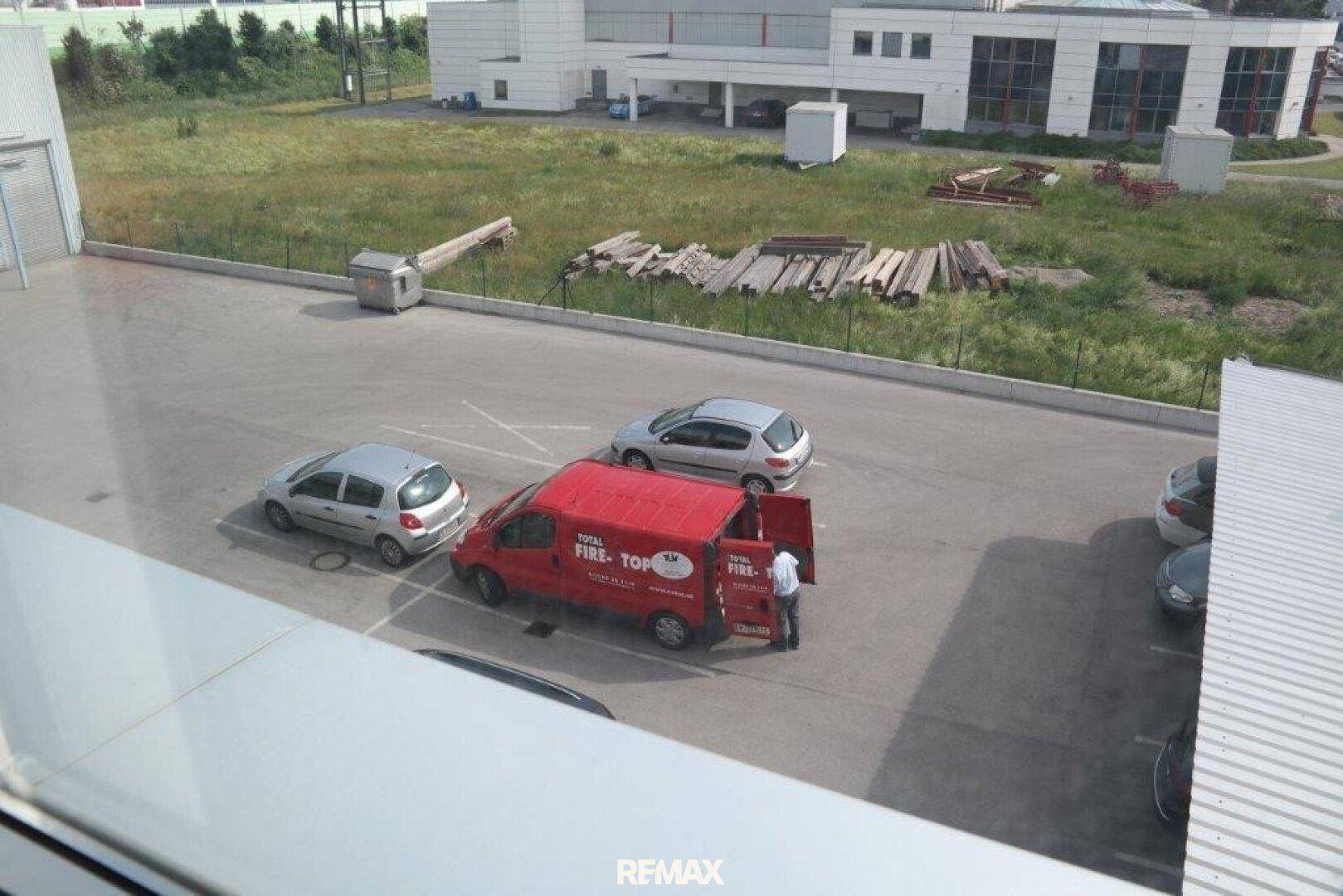 Blick aus dem Fenster auf einen Parkplatz und umliegende Industriegebäude mit Grünflächen.