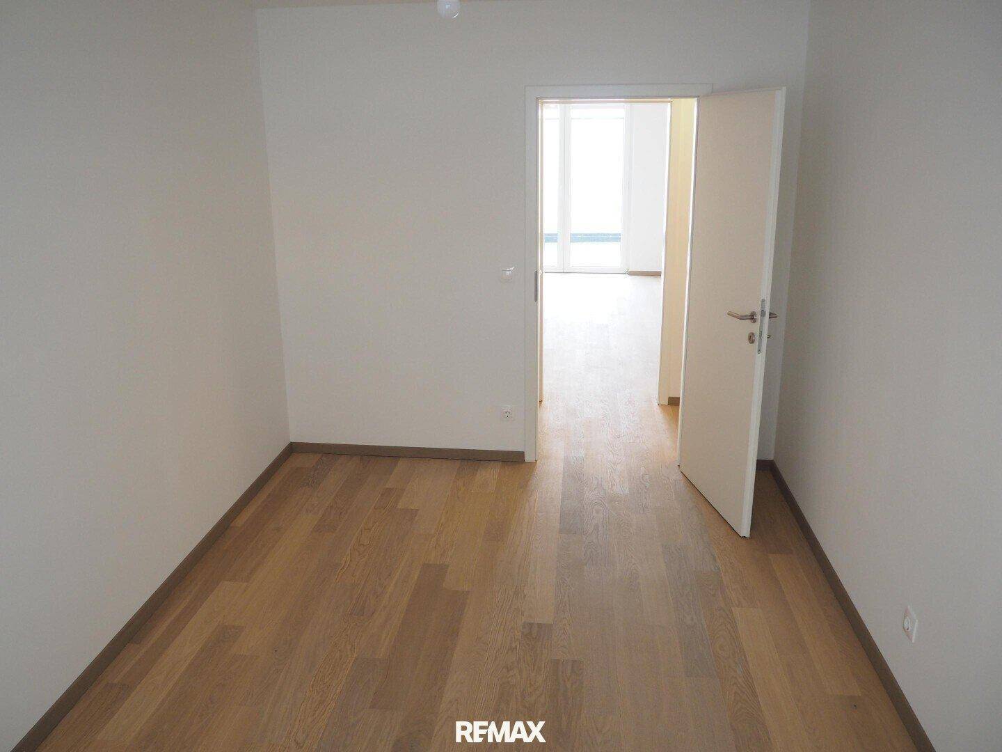 Helles Schlafzimmer mit Holzboden und offener Tür zu einem weiteren Wohnbereich.