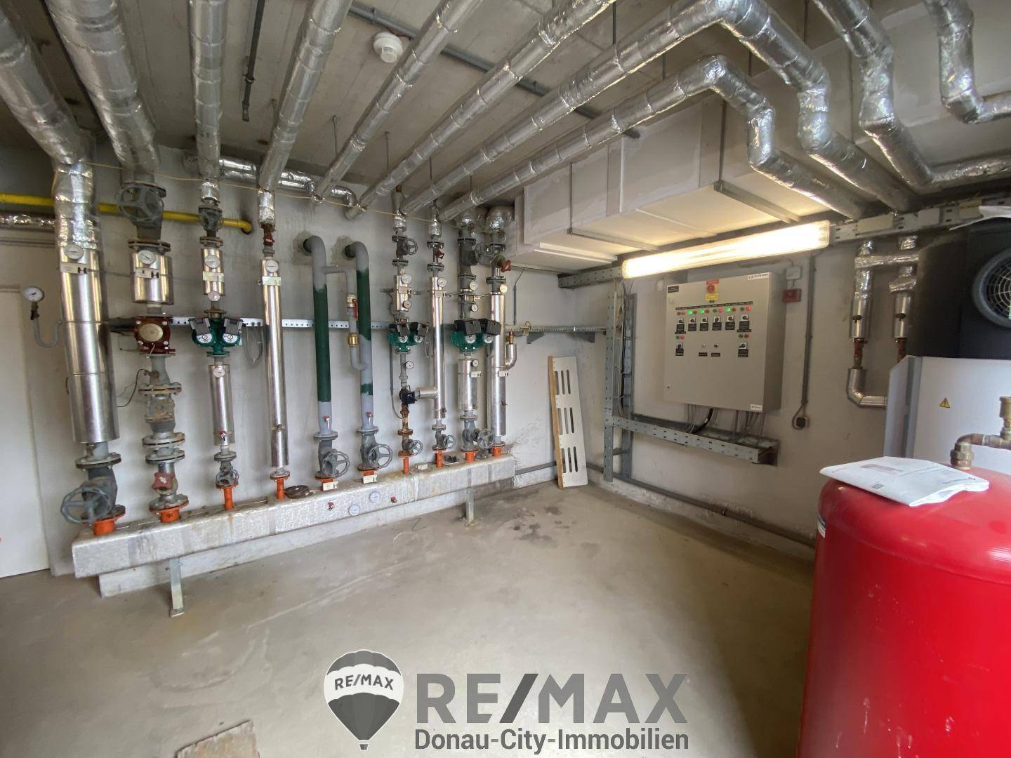 Heizungsraum mit umfangreicher Rohrinstallation, Ventilen und einem roten Warmwasserspeicher.
