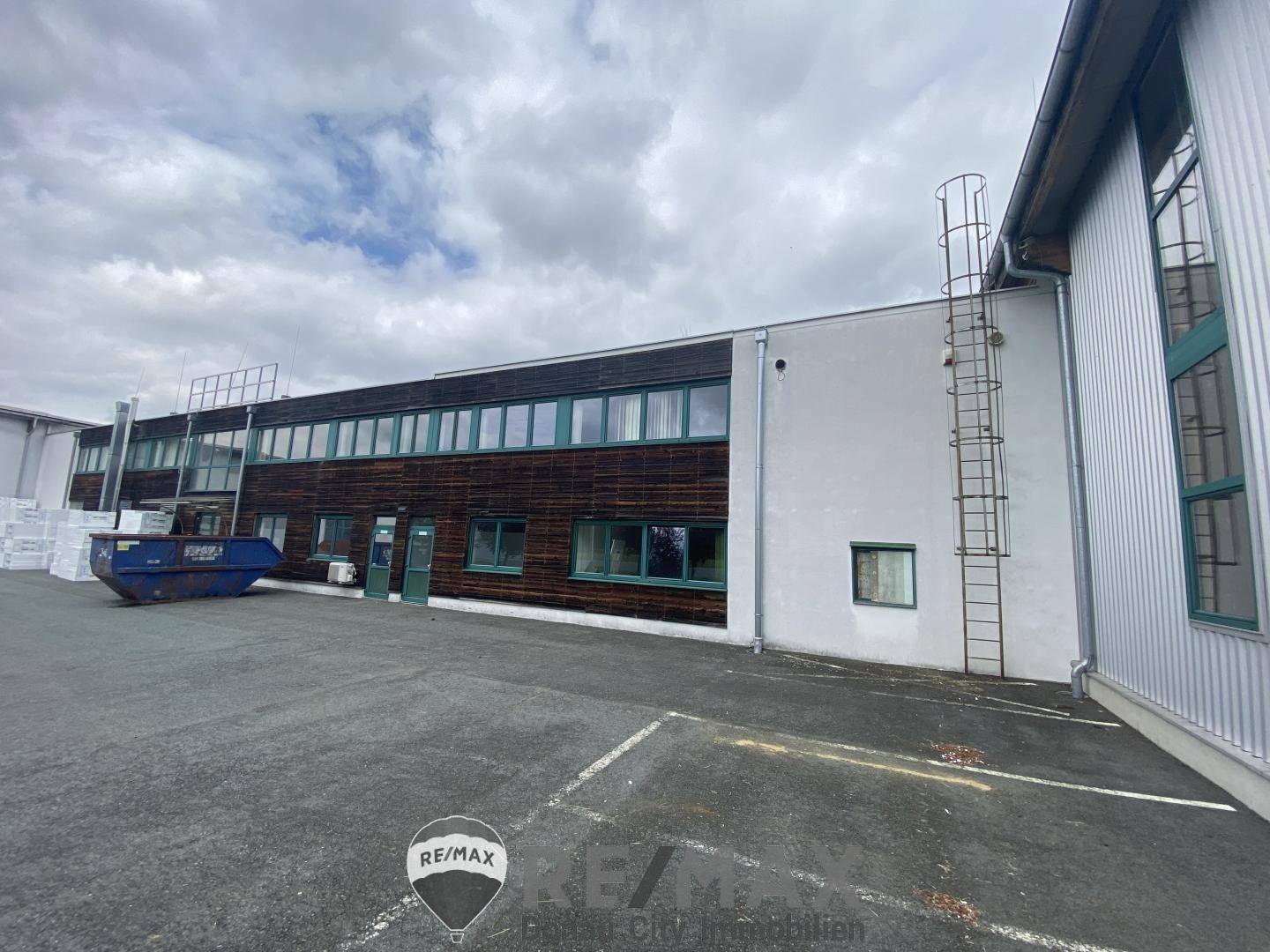 Seitenansicht eines Industriegebäudes mit Holzverkleidung und grünen Fensterrahmen, daneben ein Müllcontainer.