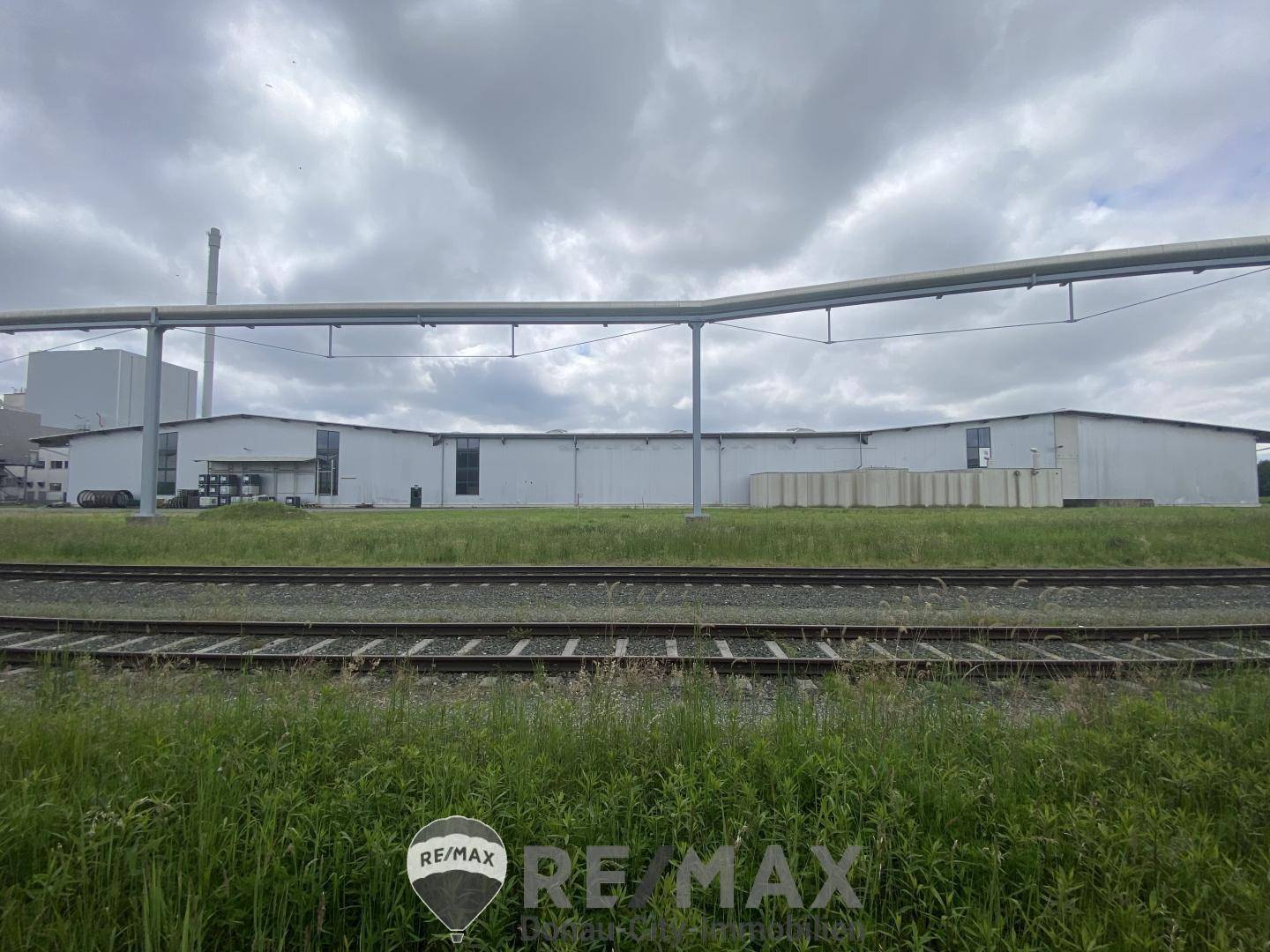 Weitläufige Ansicht eines Industriegebäudes hinter Bahngleisen und grüner Wiese unter bewölktem Himmel.