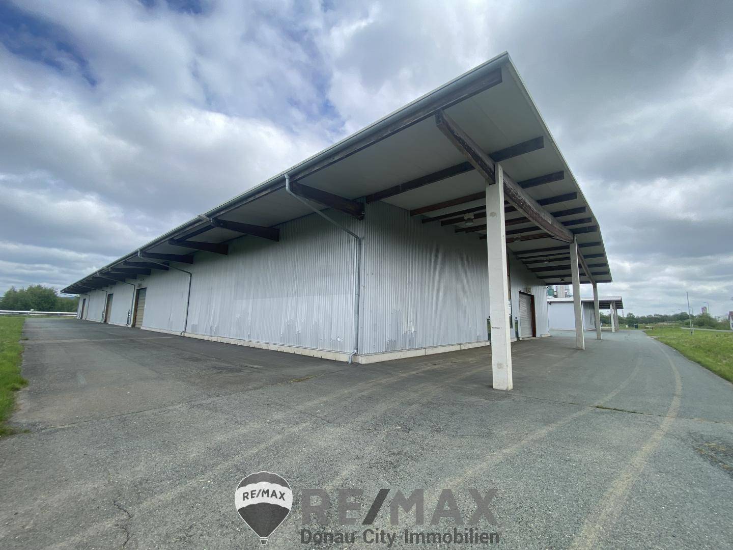 Lange Seitenfassade eines Industriegebäudes mit durchgehendem Vordach und breitem Zufahrtsweg unter bewölktem Himmel.
