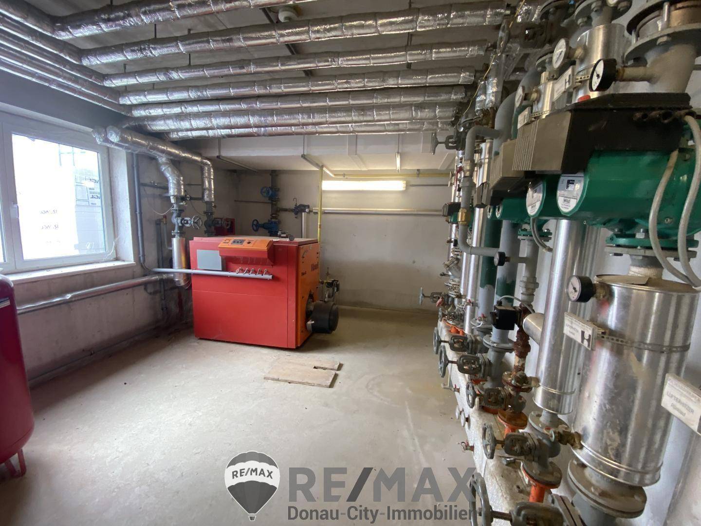 Heizungsraum mit einem großen roten Heizkessel, vielen Rohren und einem Fenster, das Tageslicht hereinlässt.