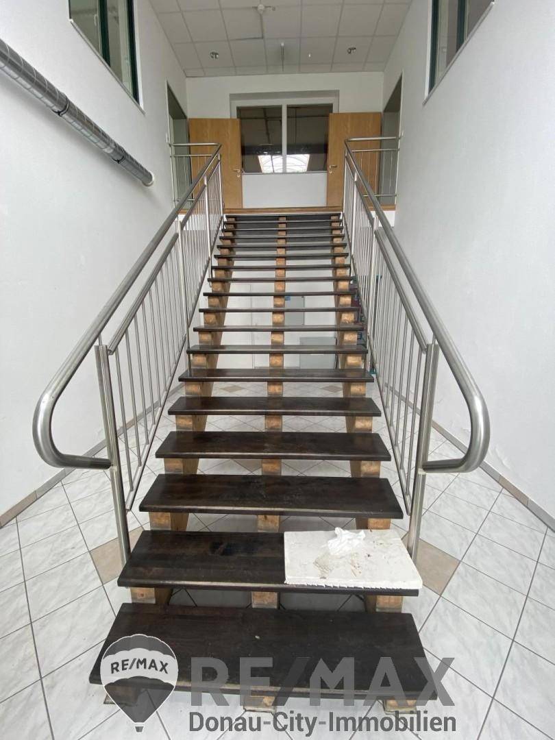 Treppenhaus mit Holzstufen, Edelstahlgeländer und Fliesenboden, führt zu oberen Etagen.