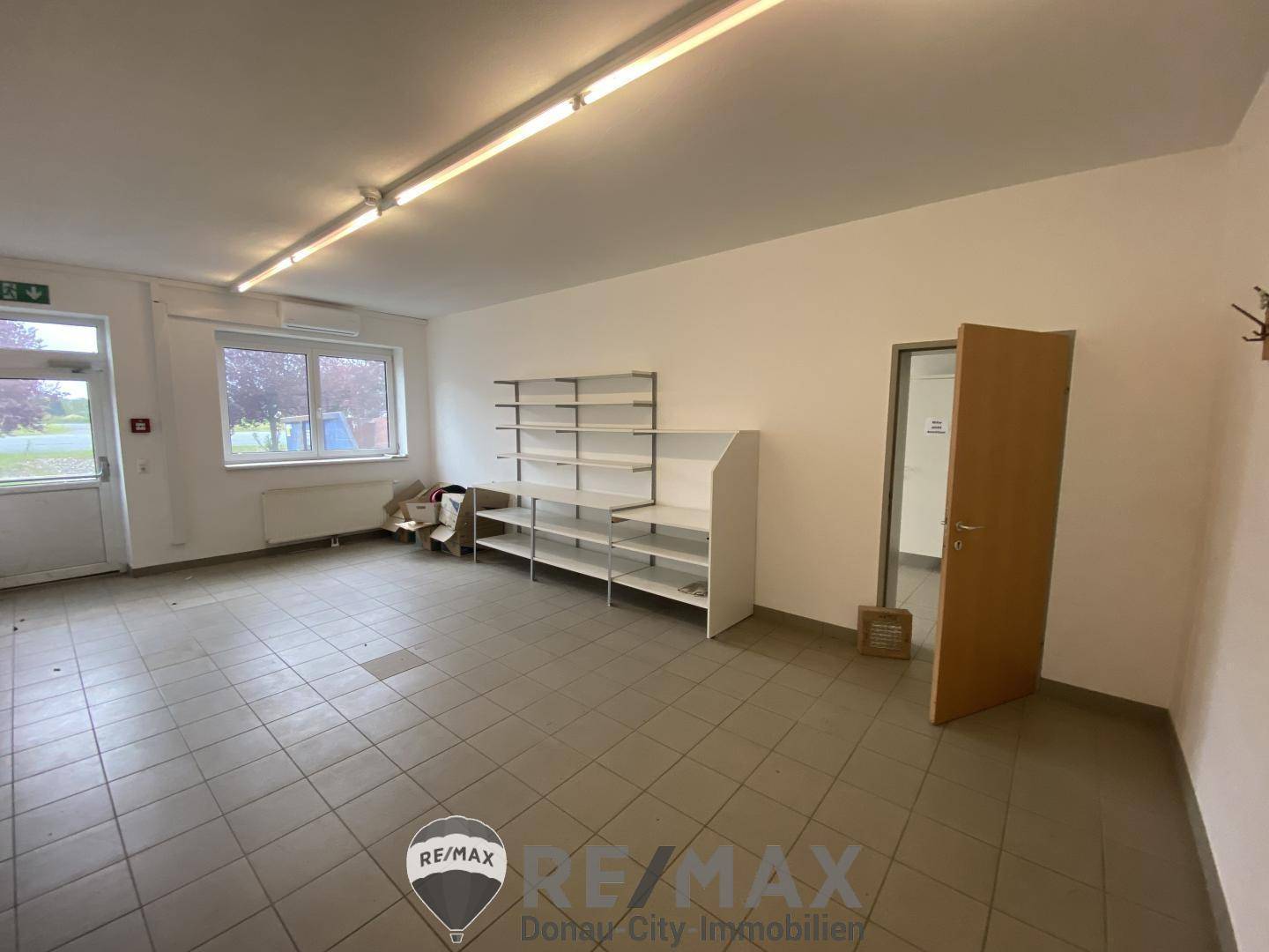 Leerer Raum mit Regalen, Fliesenboden und einem Fenster, das Tageslicht hereinlässt.