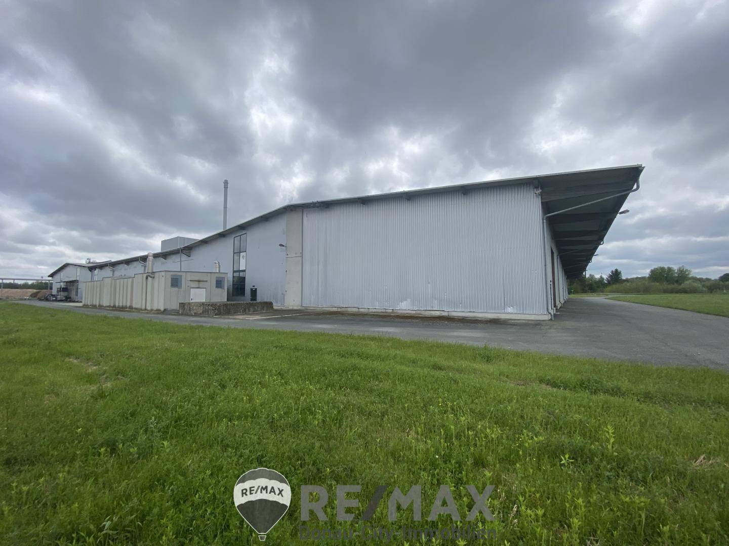 Seitenansicht des Industriegebäudes mit weitläufiger Grünfläche und grauer Wellblechfassade unter bewölktem Himmel.