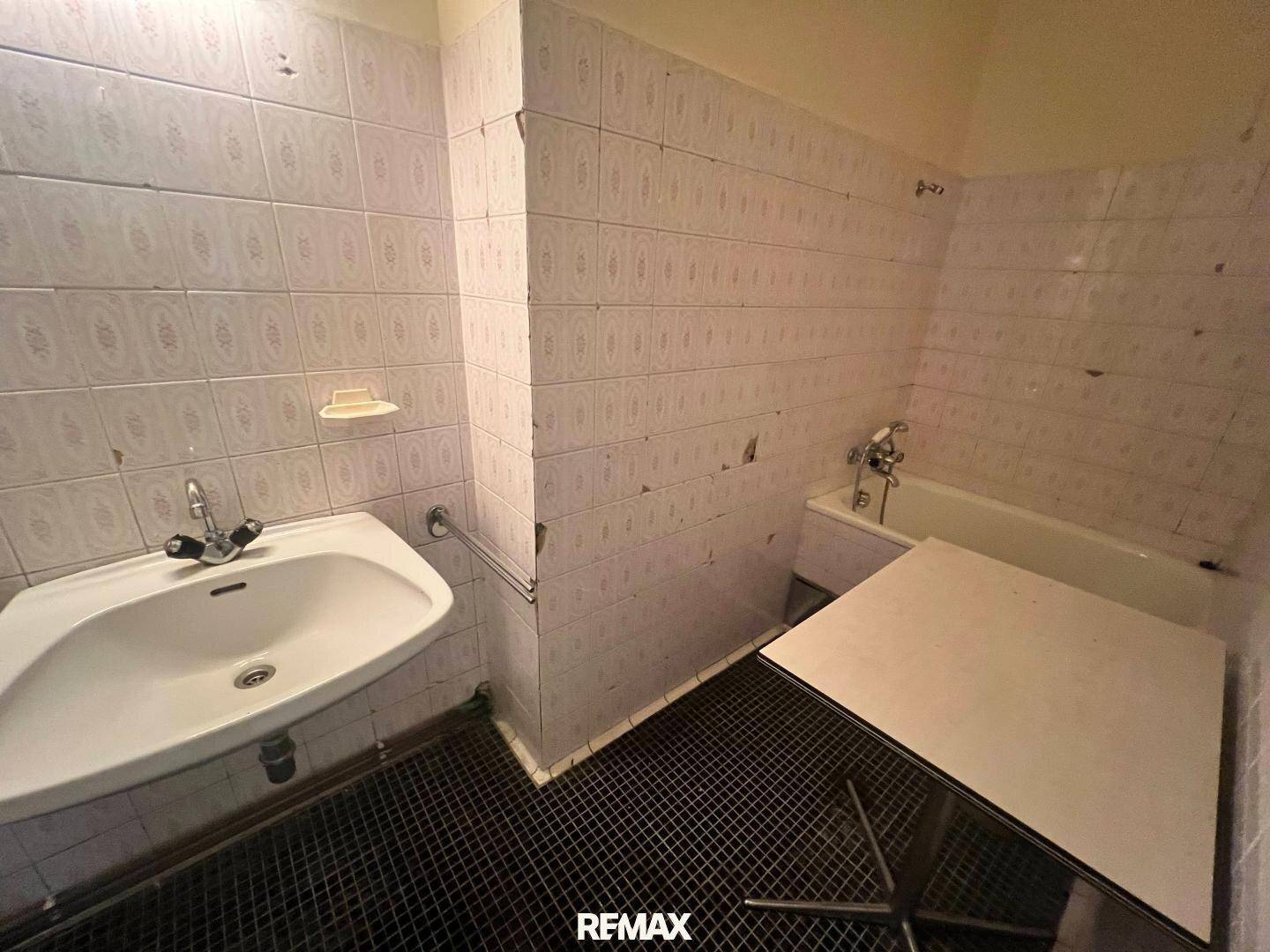 Badezimmer mit weißen Fliesen, Waschbecken und Badewanne, das einen renovierungsbedürftigen Zustand aufweist.