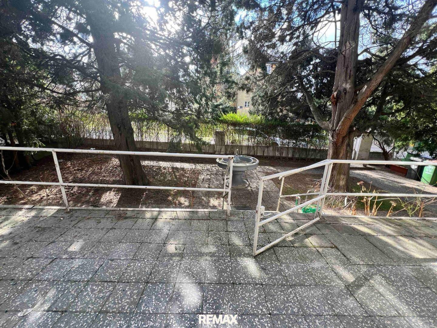 Kleine gepflasterte Terrasse mit Metallgeländer und Blick auf Bäume und einen Brunnen im Garten.