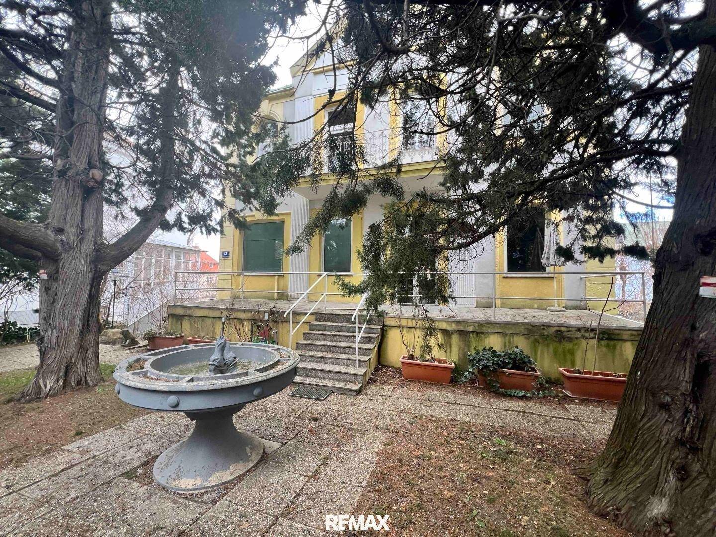 Vorderansicht eines historischen Gebäudes mit gelber Fassade, Treppe und einem dekorativen Brunnen im Vorgarten.