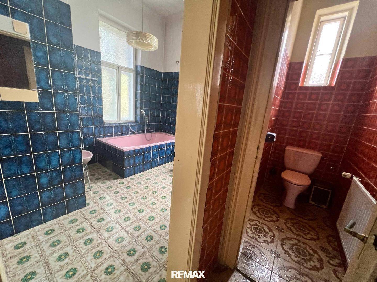 Badezimmer mit blauen Fliesen und rosa Badewanne, angrenzend an eine separate Toilette mit roten Fliesen.