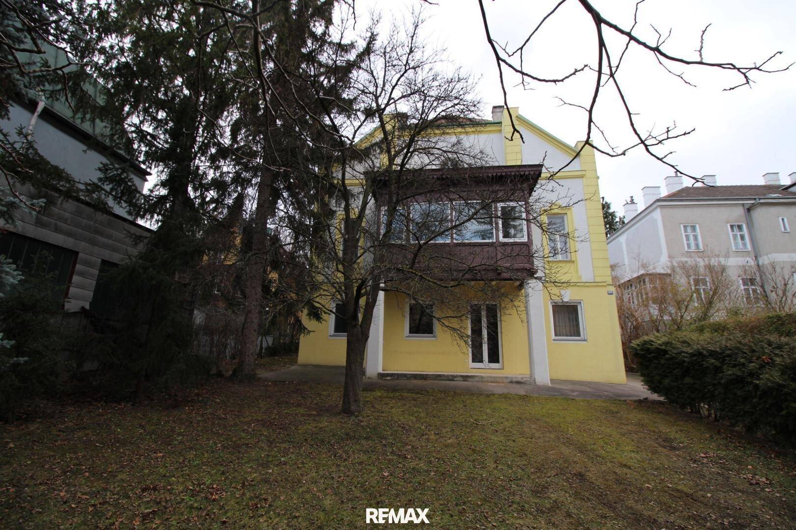 Gartenbereich hinter dem Haus mit Bäumen und einem verglasten Balkon im ersten Stock.