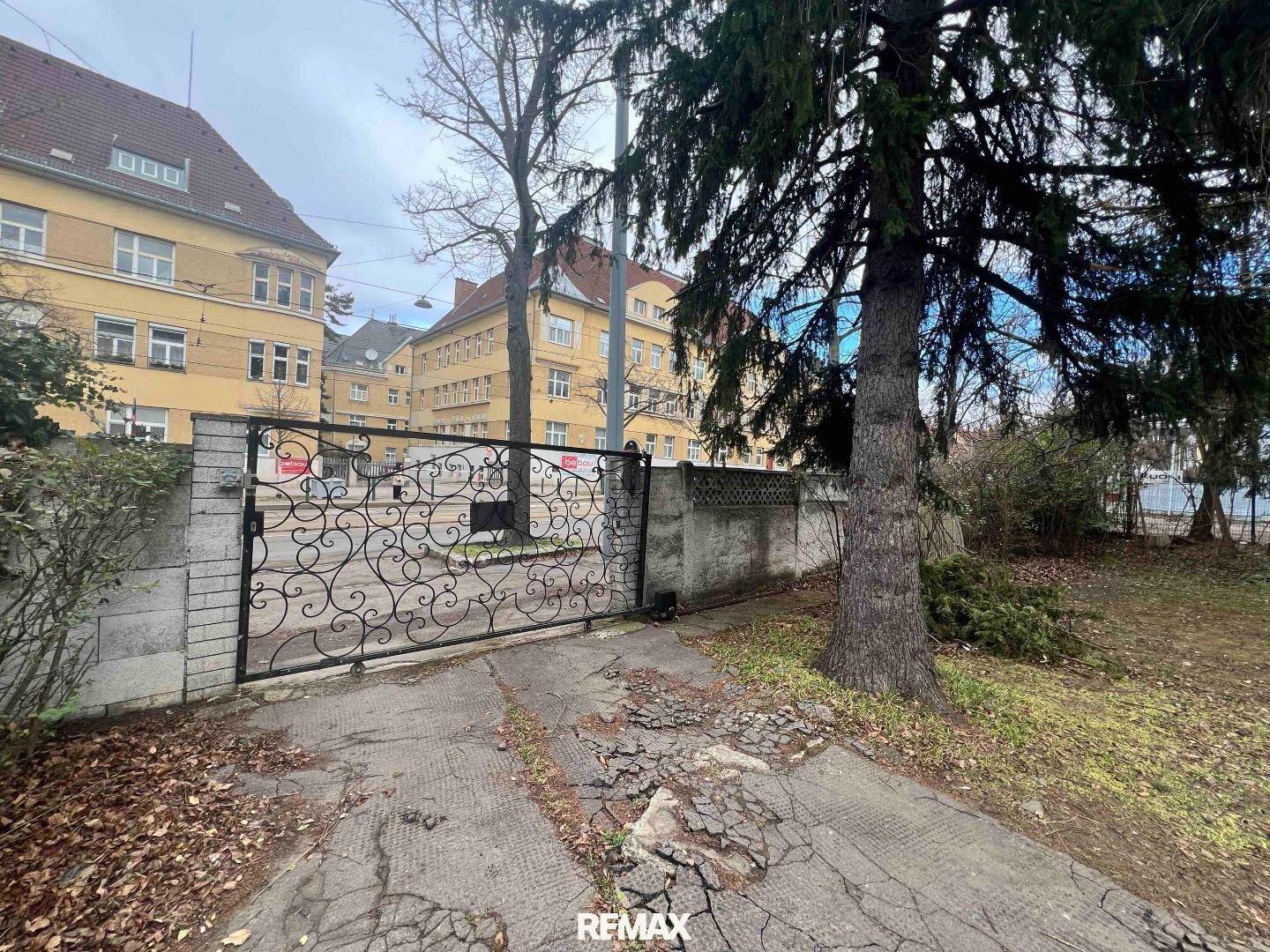 Historisches schmiedeeisernes Tor mit Zufahrt zu einem Gebäude, umgeben von Bäumen und weiteren Häusern.