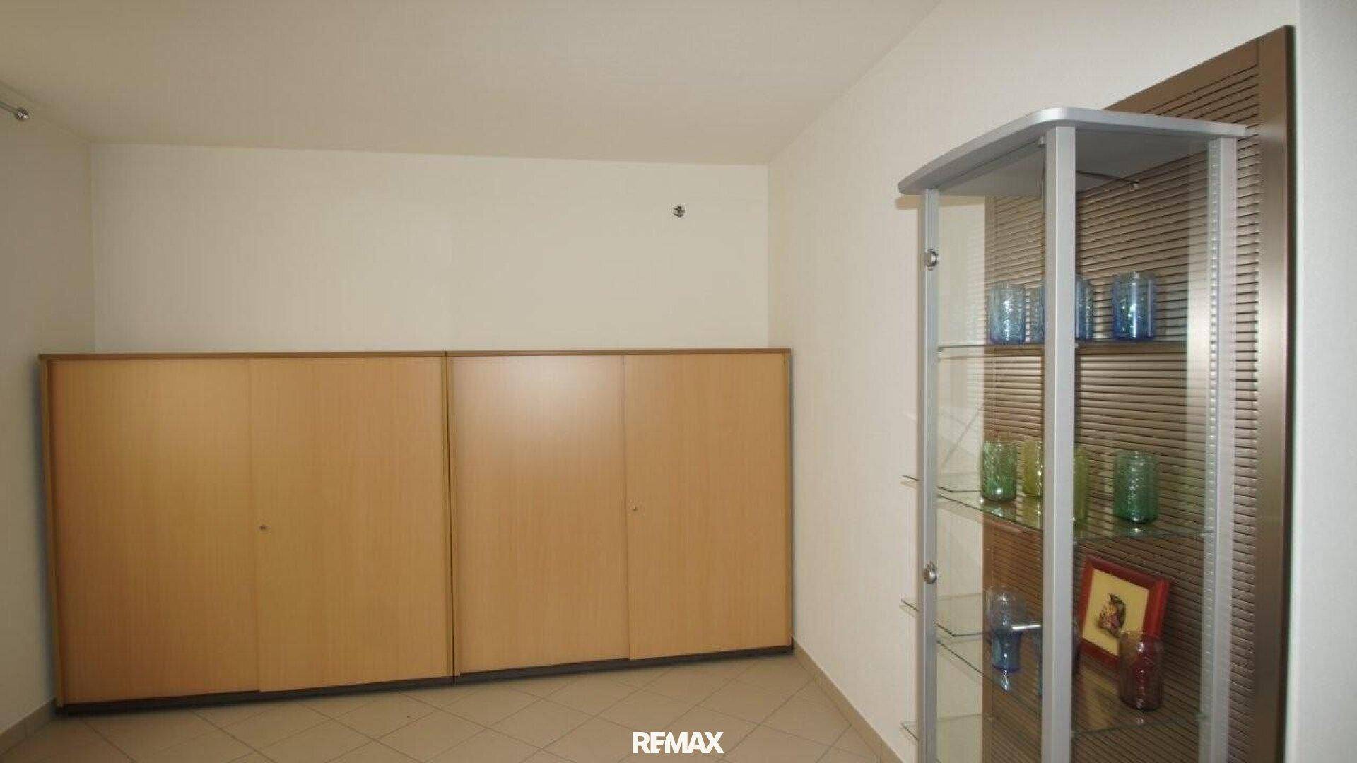 Raum mit Holzschränken, einer Glasvitrine und Fliesenboden, ideal für Lagerung oder Büro.