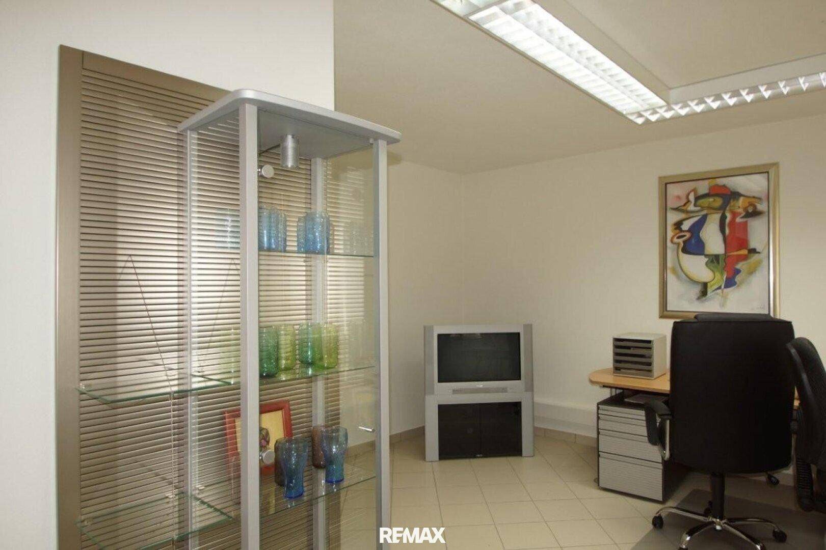 Helles Büro mit Glasvitrine, Schreibtisch und einem farbenfrohen Gemälde.