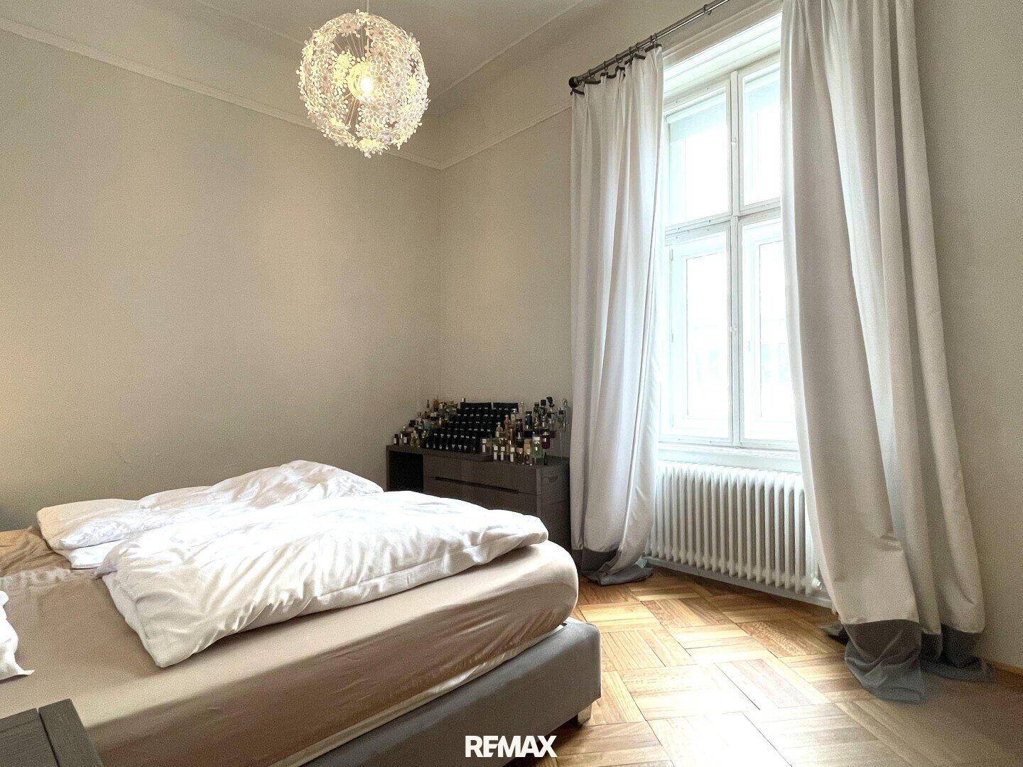Helles Schlafzimmer mit Bett, großem Fenster und weißen Vorhängen, Parkettboden.