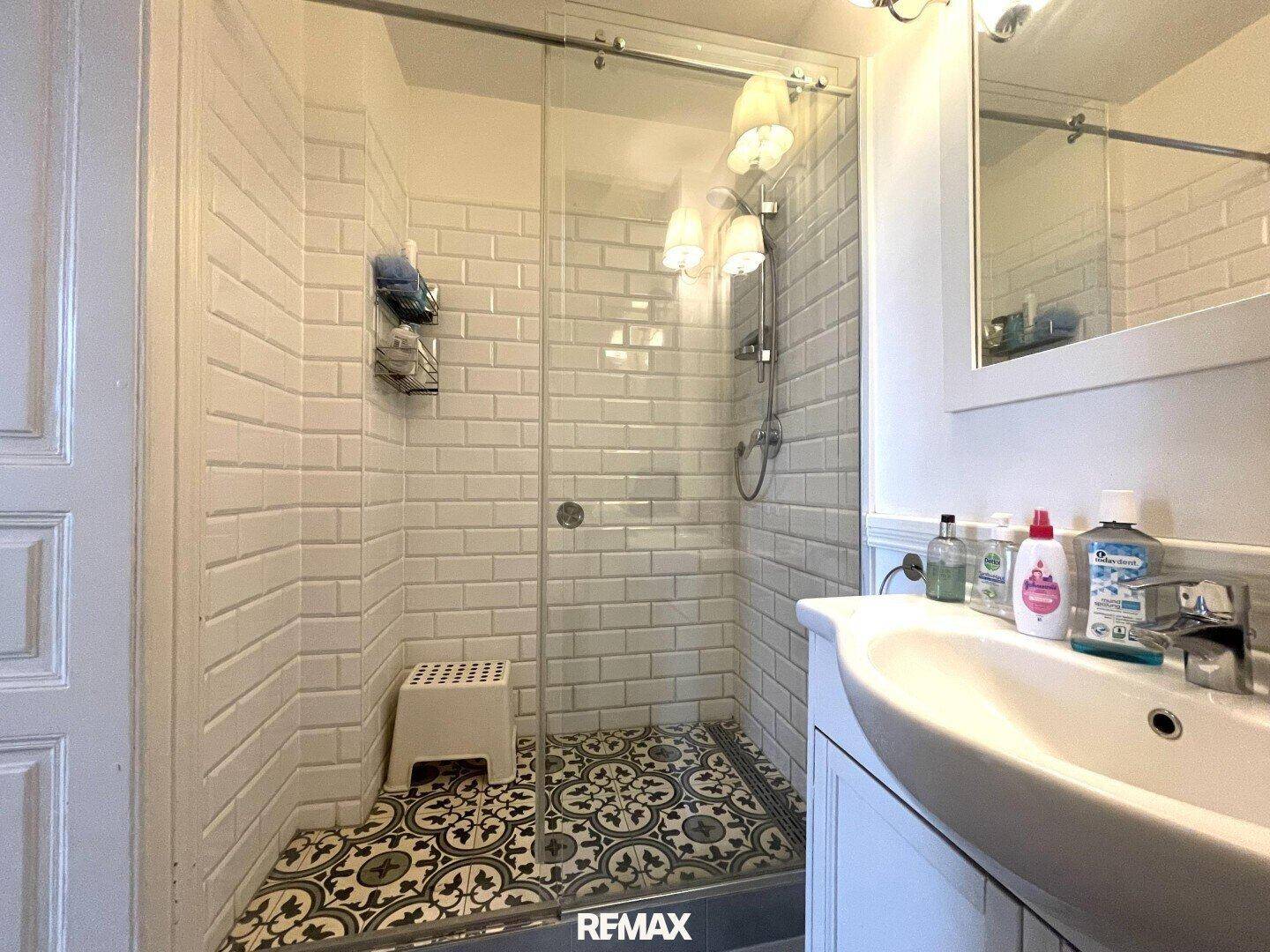 Duschbereich im Badezimmer mit Glasabtrennung, weißen Fliesen und dekorativem Bodenmuster.