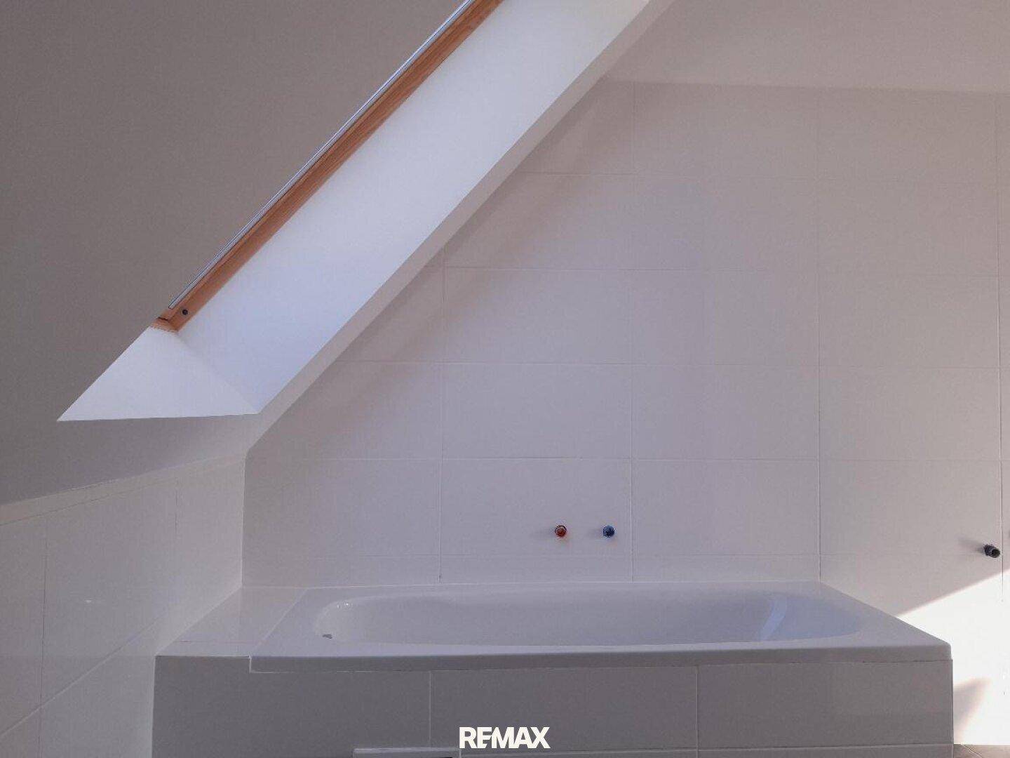 Helles Badezimmer im Dachgeschoss mit Badewanne, weißen Fliesen und einem Dachfenster.