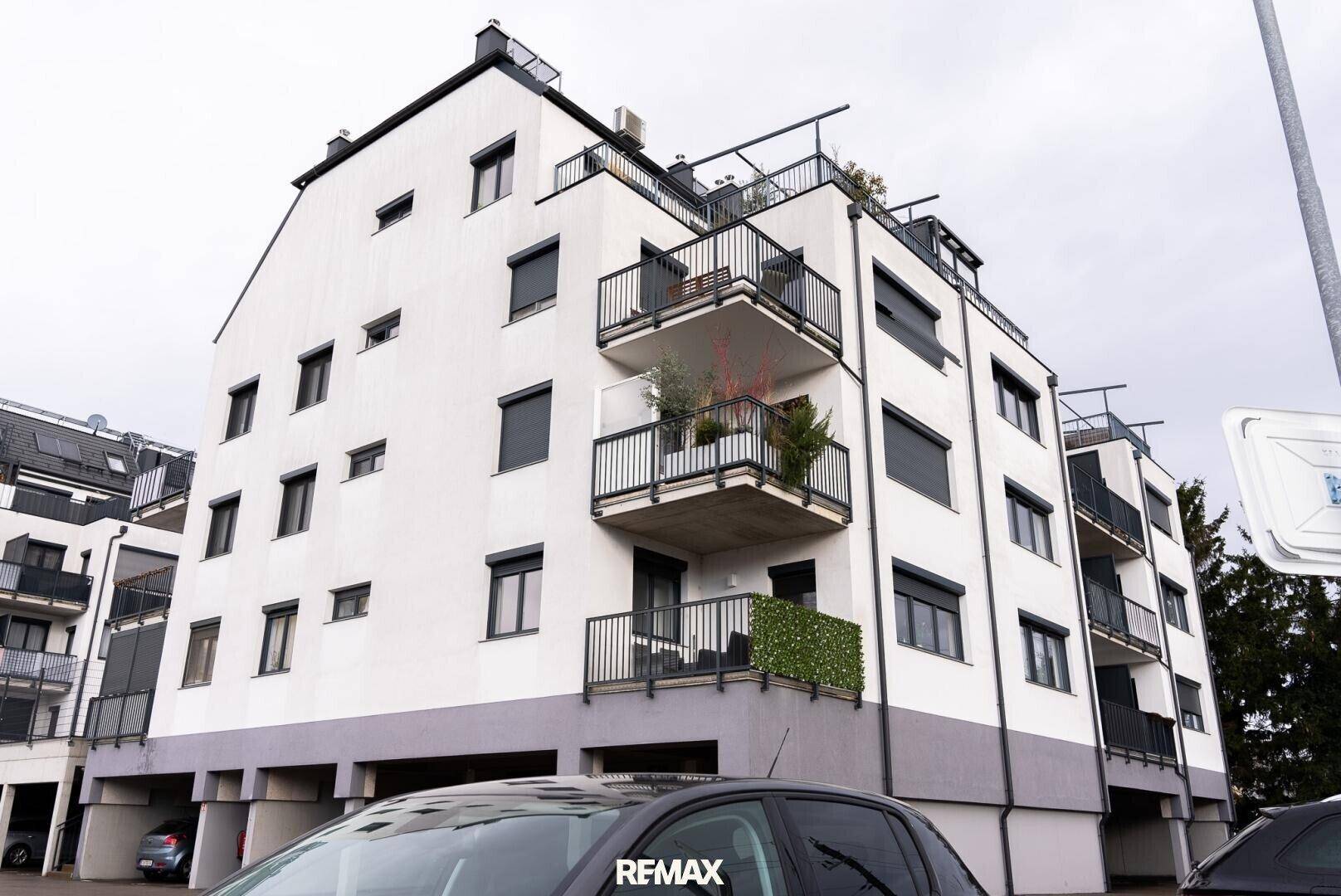 Modernes Wohngebäude mit weißen Wänden, dunklen Balkongeländern und einigen Pflanzen auf den Balkonen.
