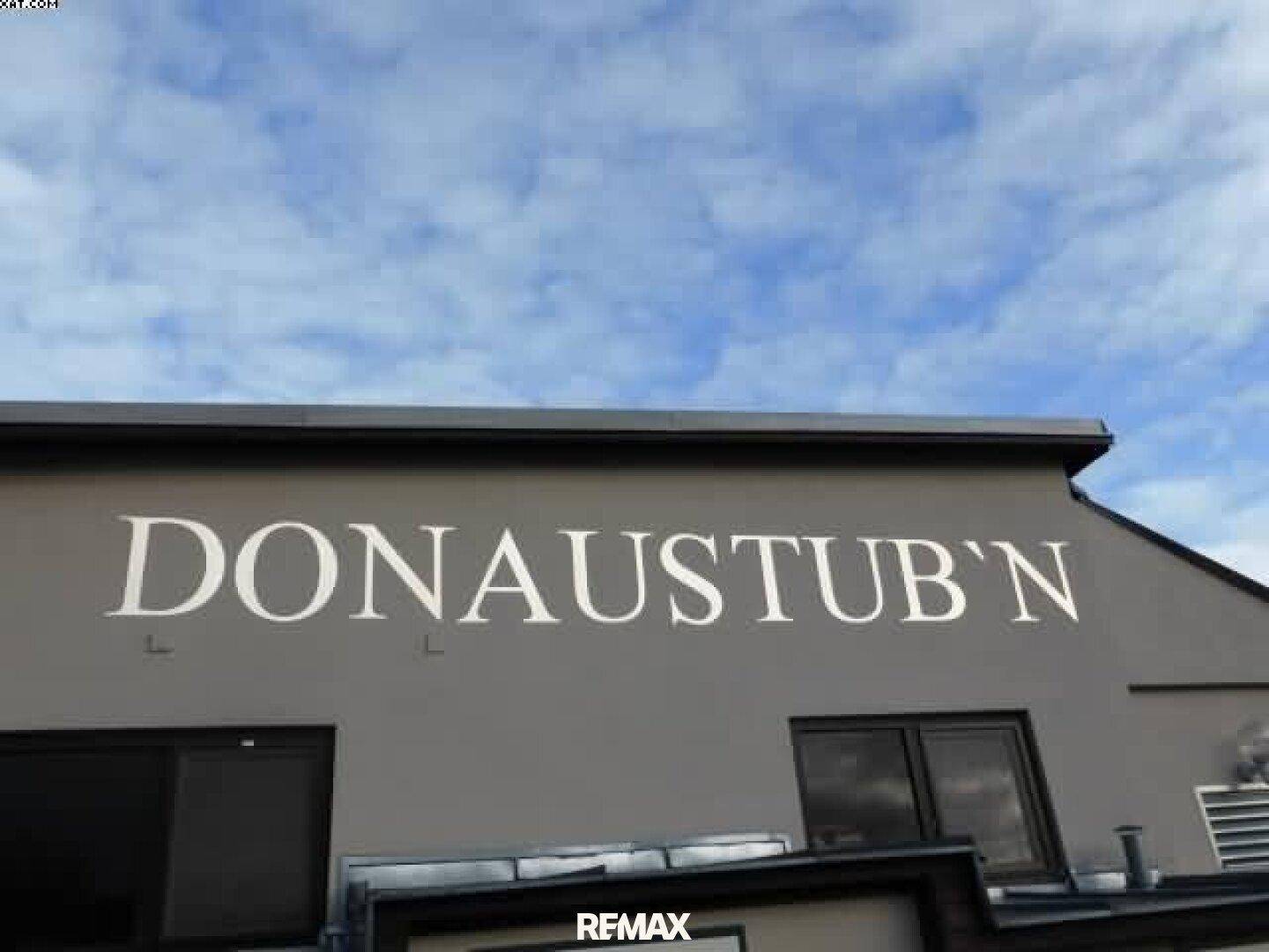 Nahaufnahme der Gebäudefassade mit dem Schriftzug "DONAUSTUB'N" unter einem bewölkten Himmel.