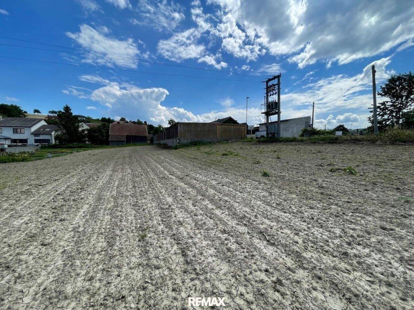 Weitläufiges Ackerland unter blauem Himmel mit ländlicher Bebauung im Hintergrund.