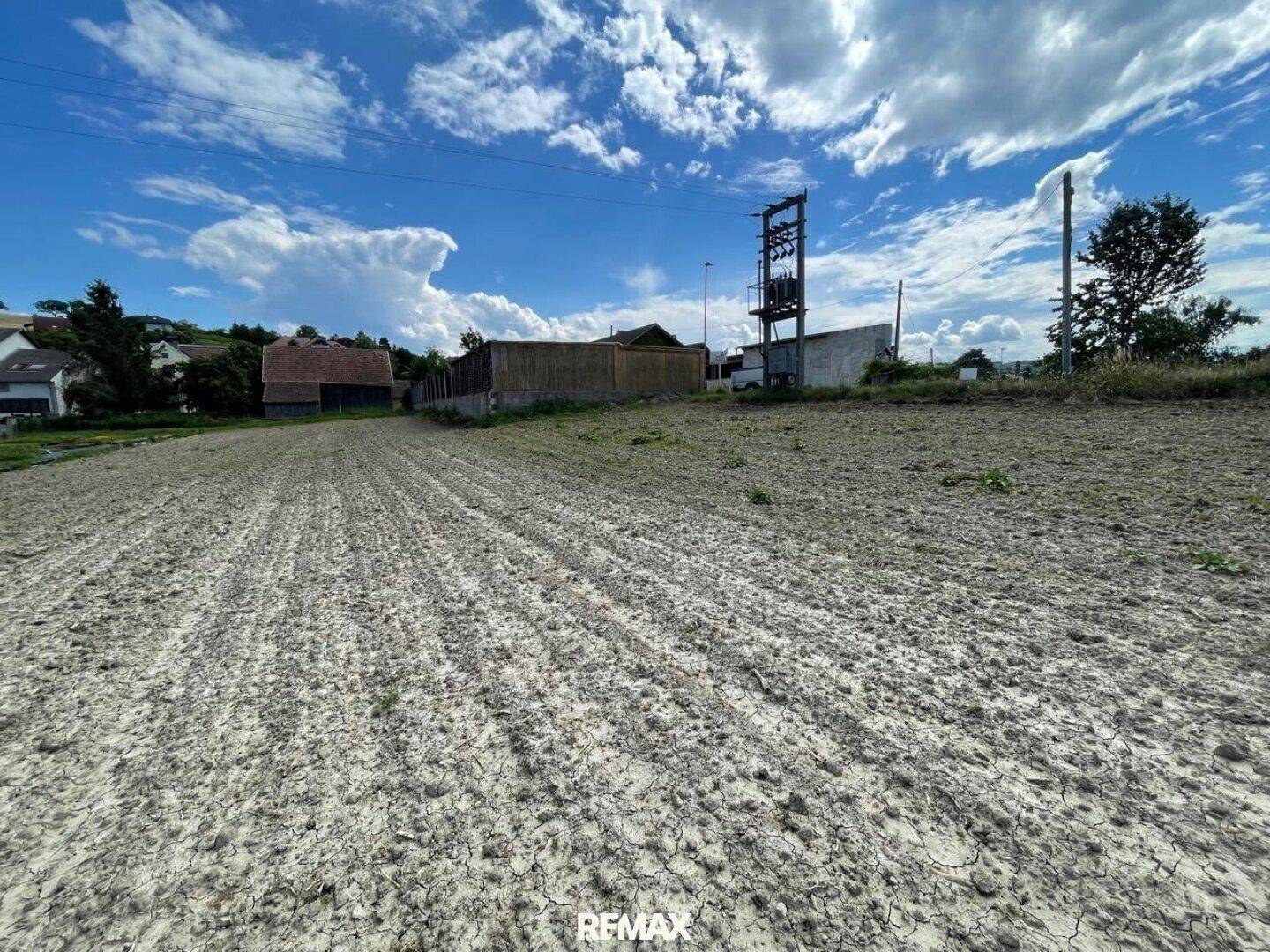 Offenes Feld mit trockener Erde und Strommast vor bewölktem Himmel.