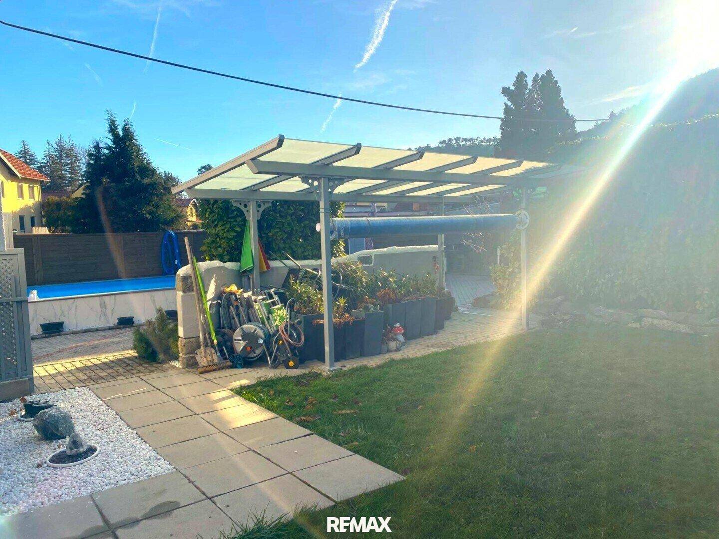 Außenbereich mit Carport-Struktur, angrenzendem Poolbereich und Rasenfläche unter strahlendem Himmel.