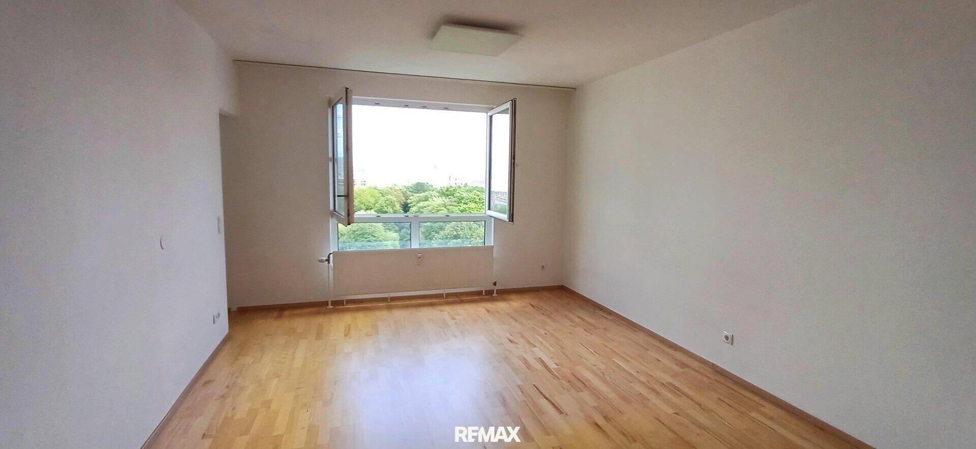 Heller Raum mit Parkettboden und Blick ins Grüne durch das geöffnete Fenster.