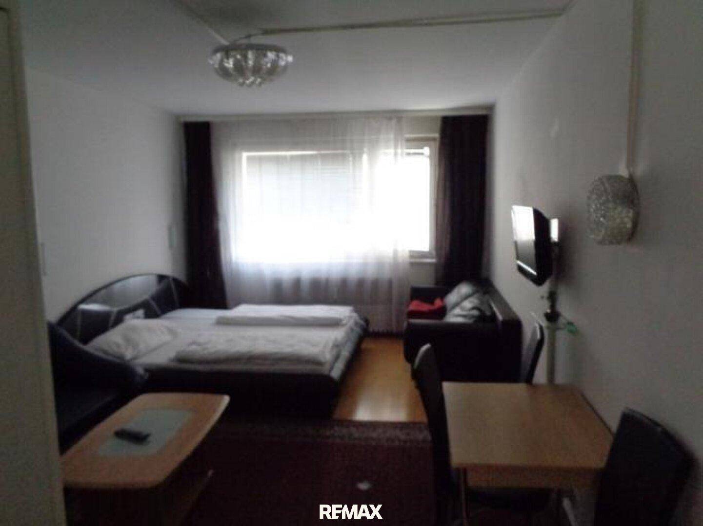 Gemütliches Wohnzimmer mit Bett, Sofa und Esstisch, beleuchtet durch ein großes Fenster.