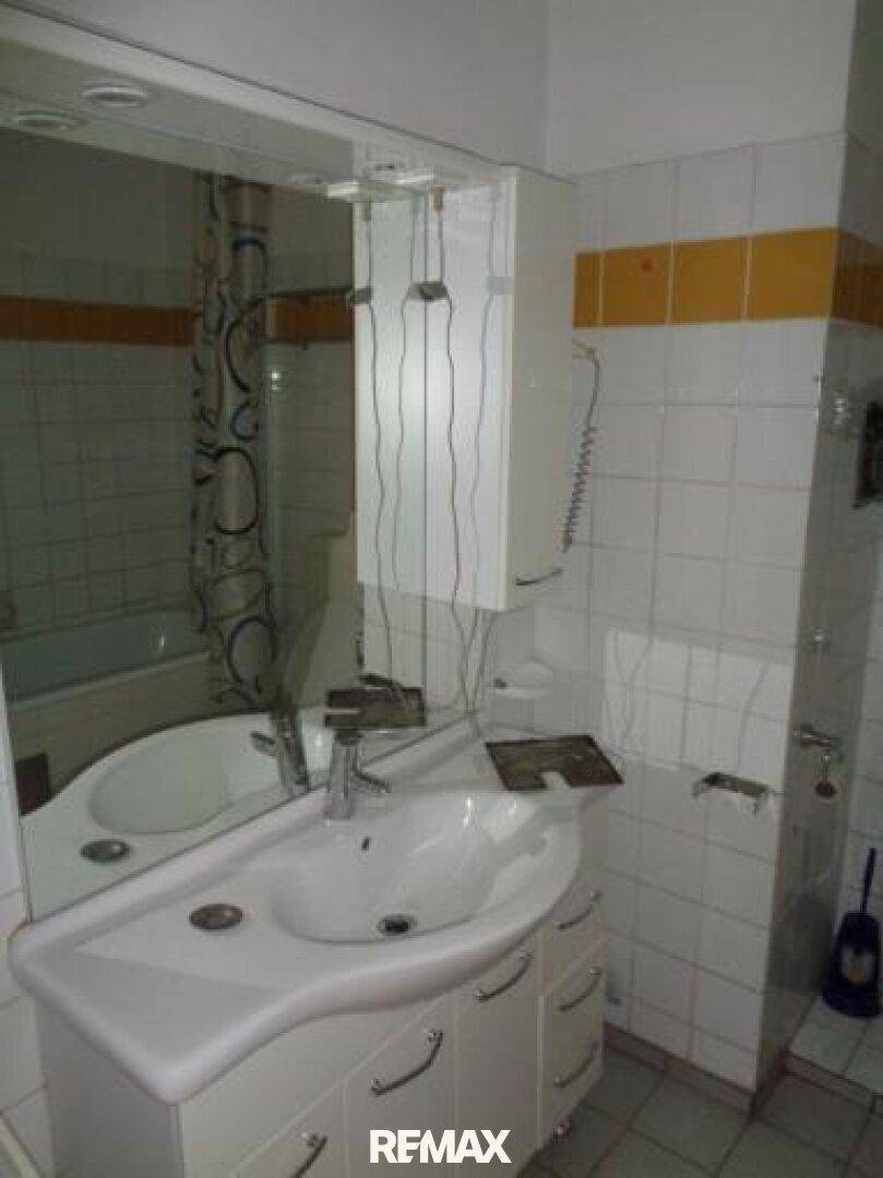 Badezimmer mit Waschbecken, großem Spiegel und weißen Fliesen mit gelbem Akzentstreifen.