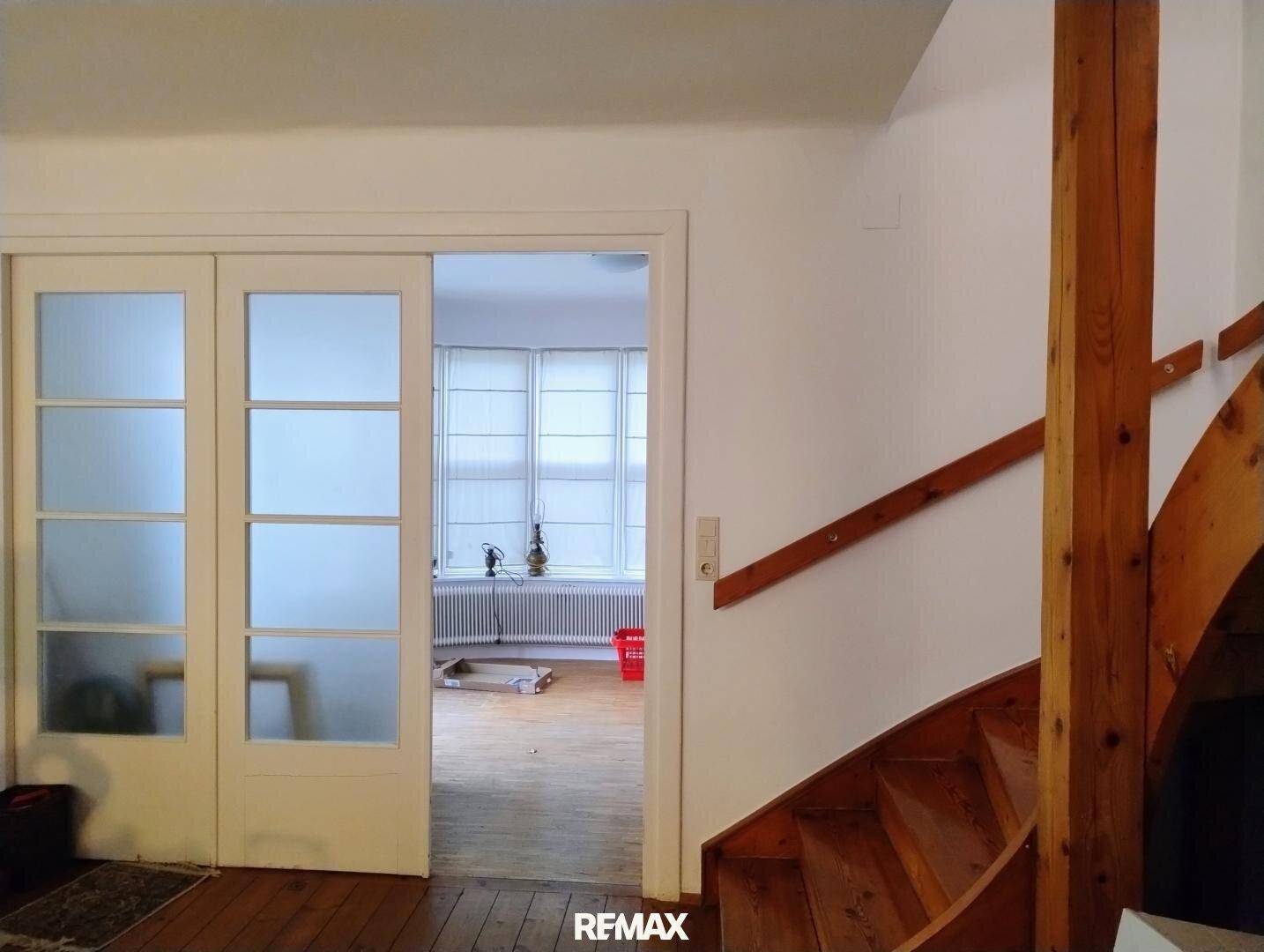 Heller Flur mit Holztreppe und Schiebetüren zu einem weiteren Raum mit Fenstern.