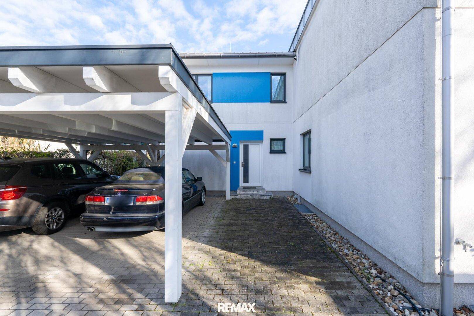 Überdachter Carport mit geparkten Autos vor dem modernen Haus unter blauem Himmel.