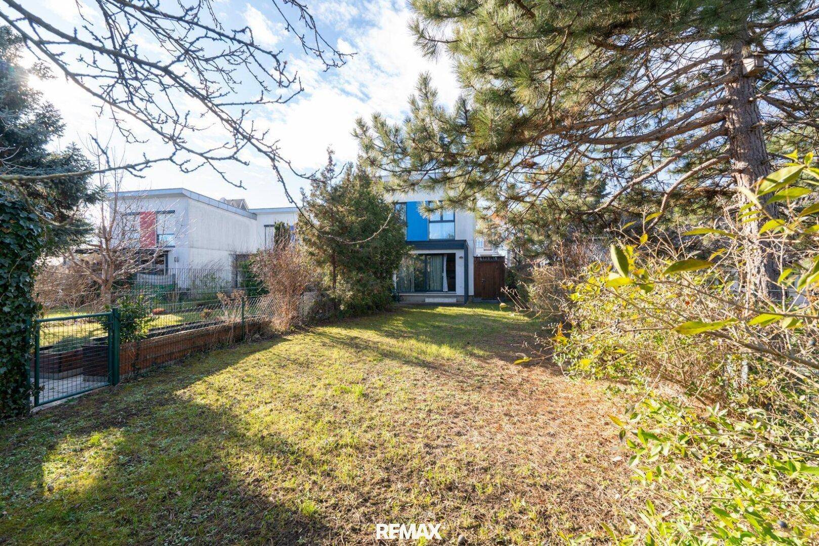 Gepflegter Garten hinter dem Haus mit Rasenfläche und hohen Bäumen unter blauem Himmel.