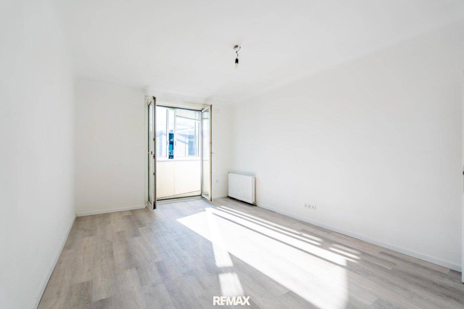 Helles, leeres Zimmer mit Laminatboden und Zugang zu einem verglasten Balkon.