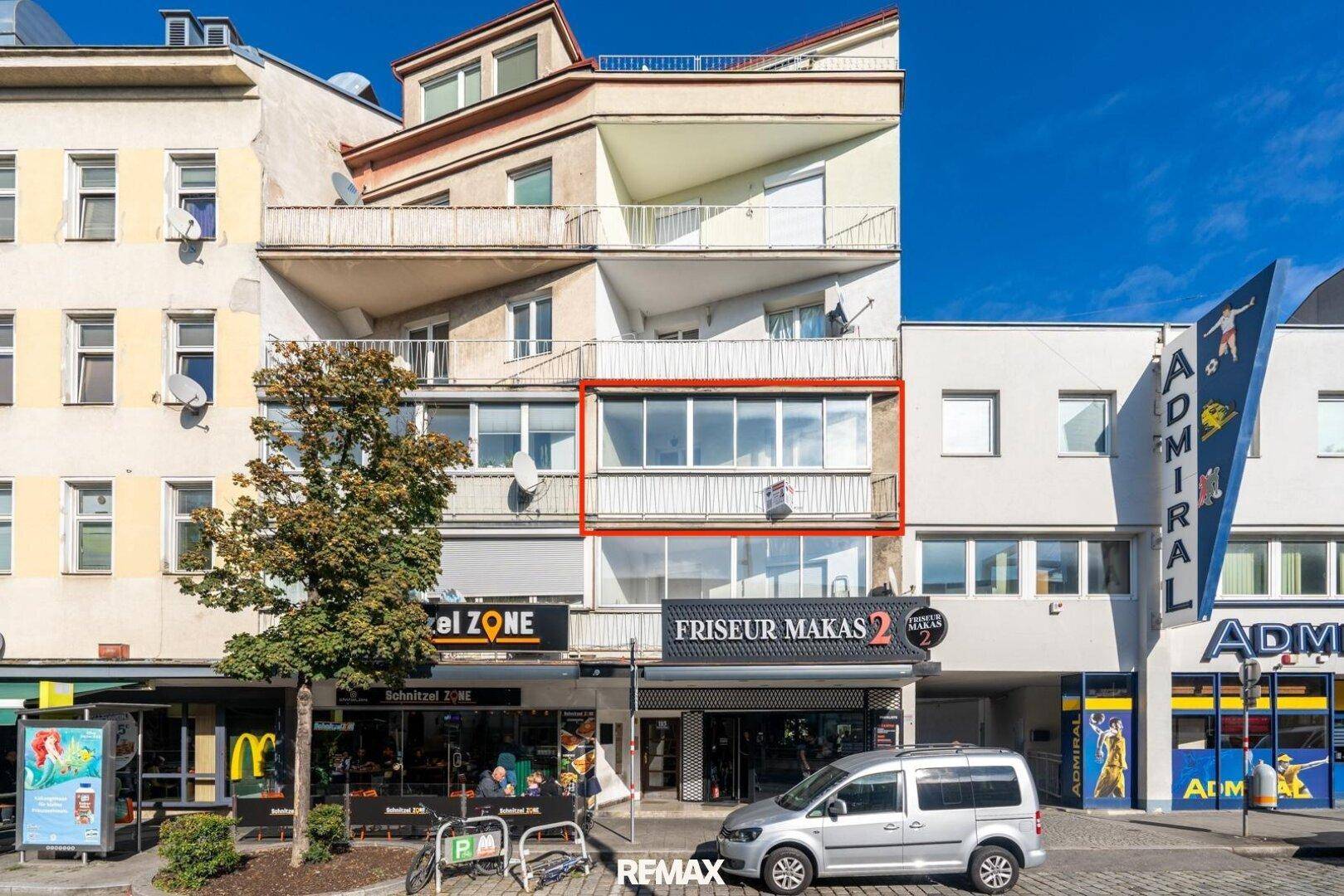 Fassade eines Mehrfamilienhauses mit Geschäften im Erdgeschoss und Wohnungen darüber in urbaner Umgebung.