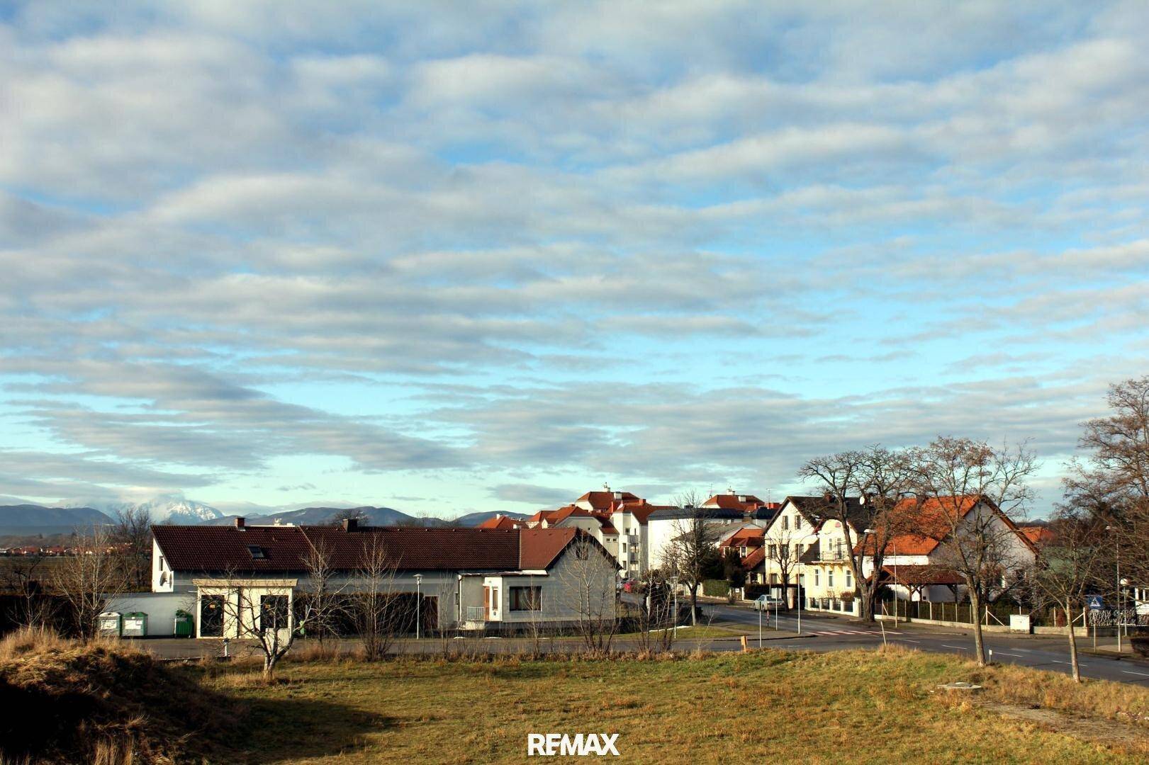 Weitläufige Landschaft mit ländlicher Bebauung und Bergen im Hintergrund unter bewölktem Himmel.
