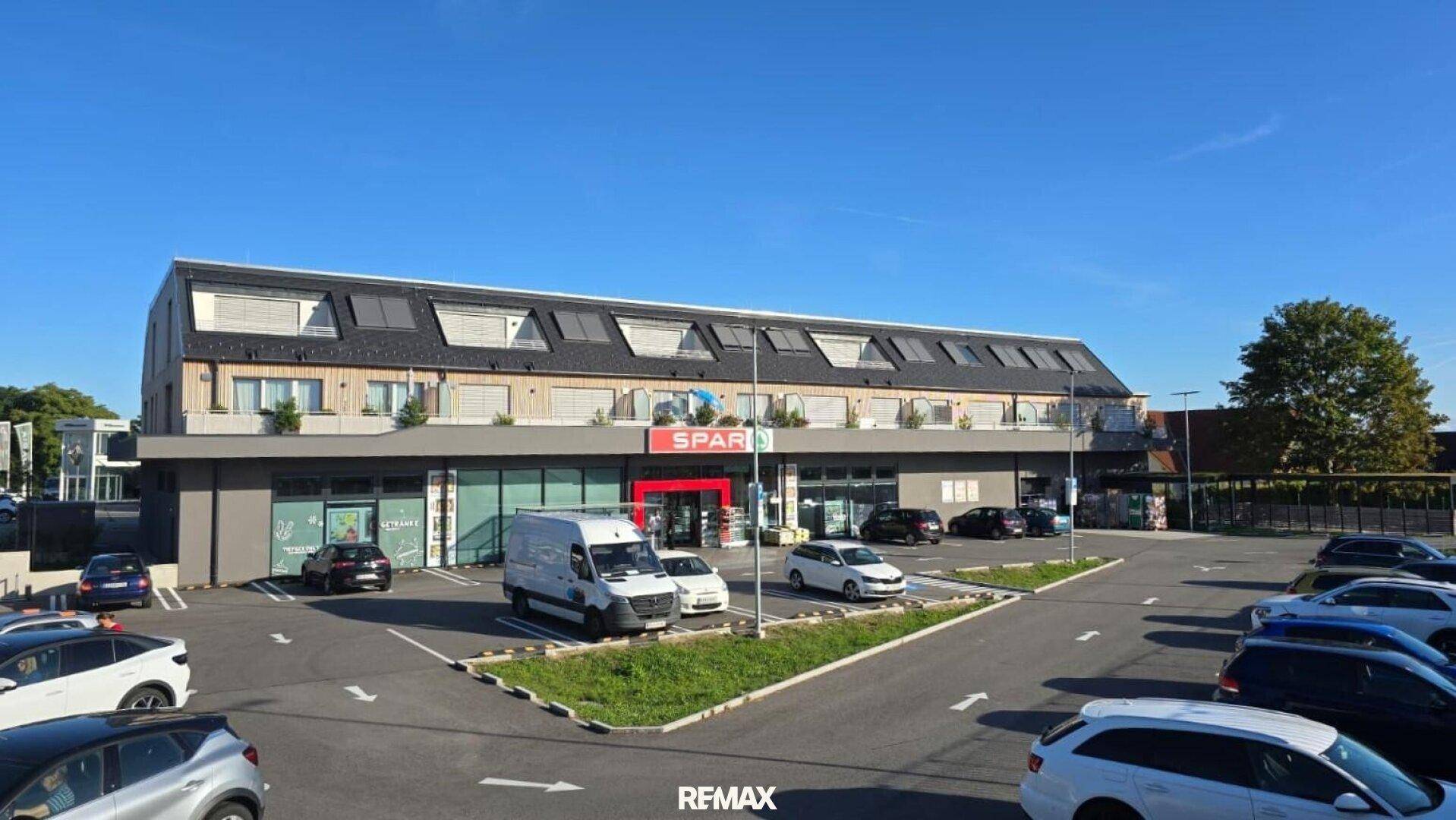Gesamtansicht des modernen Gebäudes mit Supermarkt, Wohnungen und dem dazugehörigen Parkplatz unter blauem Himmel.