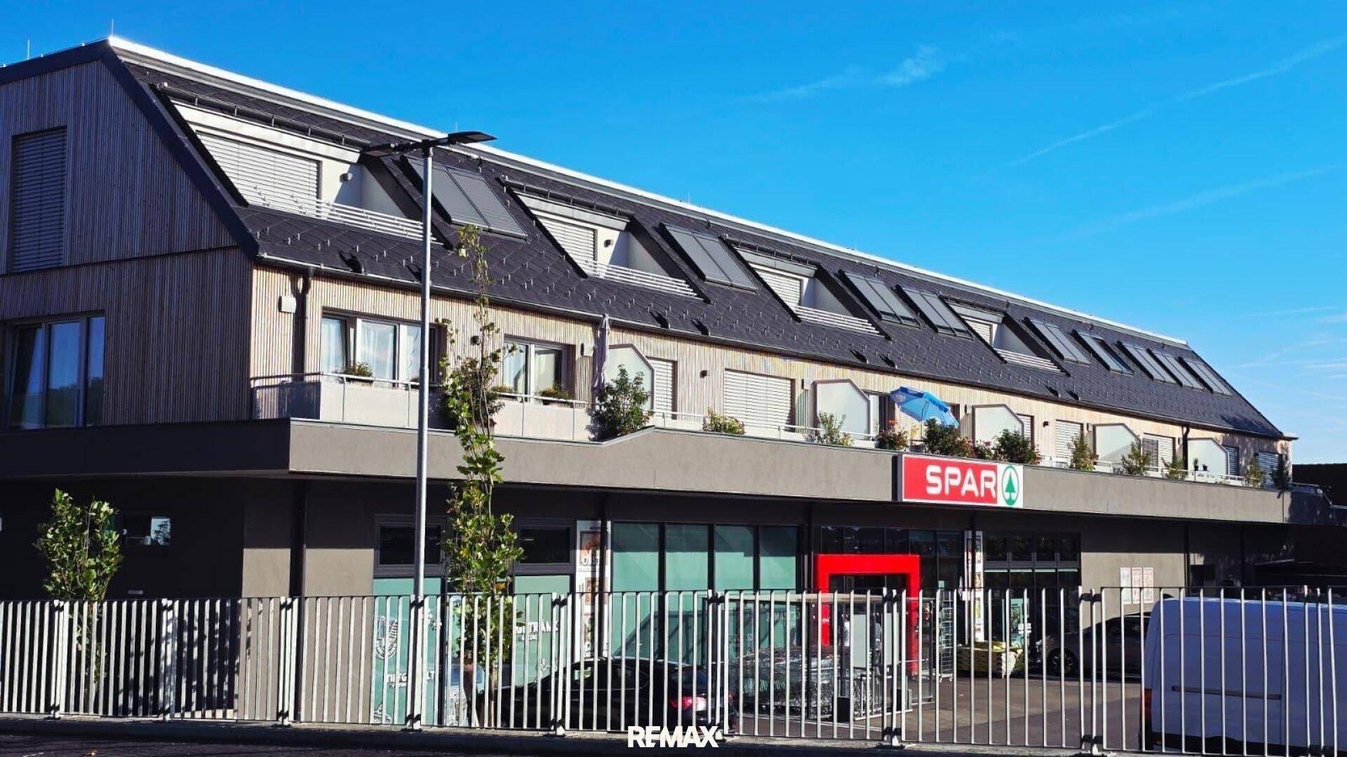 Ansicht eines modernen Gebäudes mit Supermarkt im Erdgeschoss und darüberliegenden Wohnungen unter einem Satteldach.